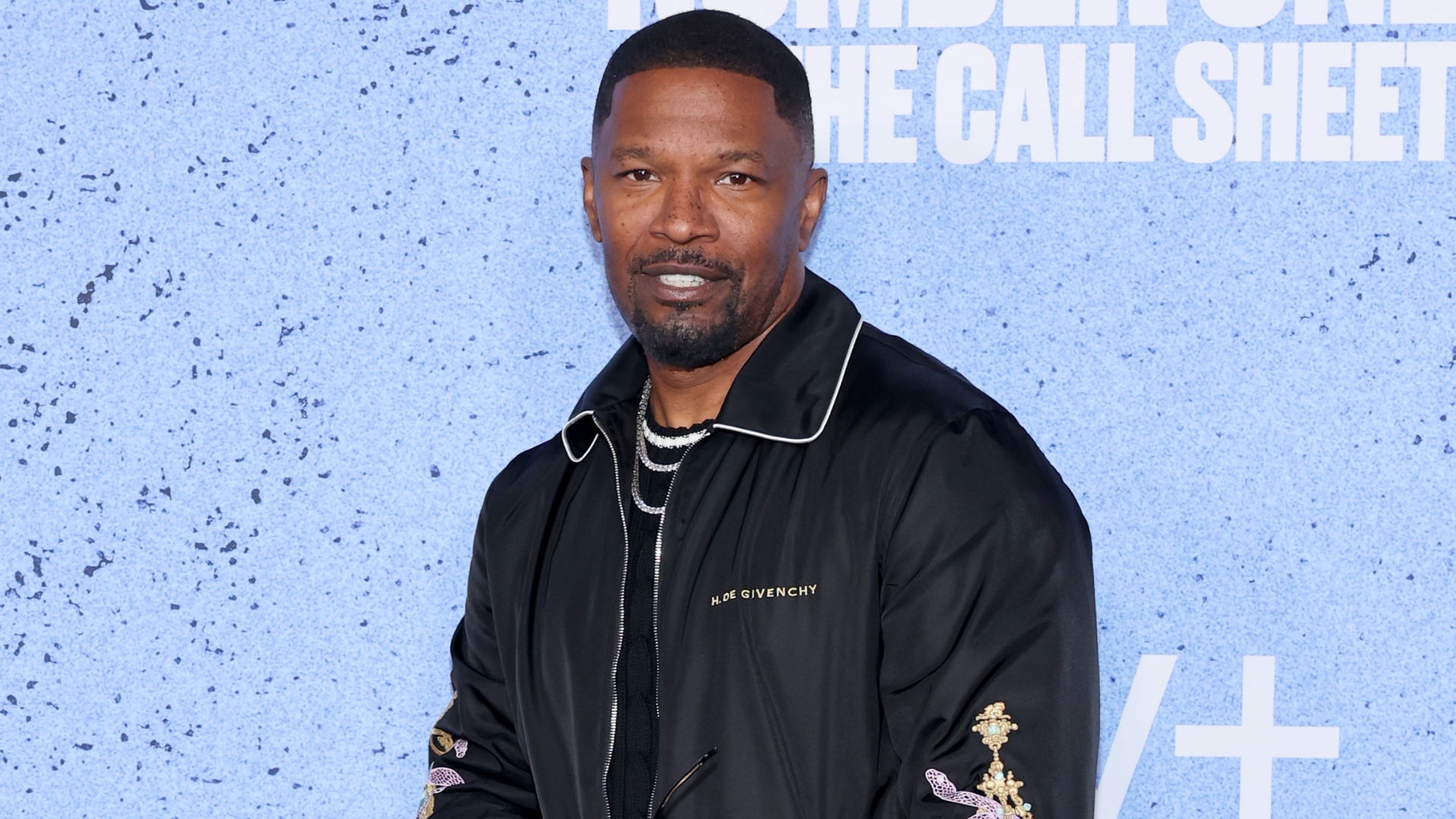 Jamie Foxx wearing a black Givenchy jacket, standing against a blue background at an event.