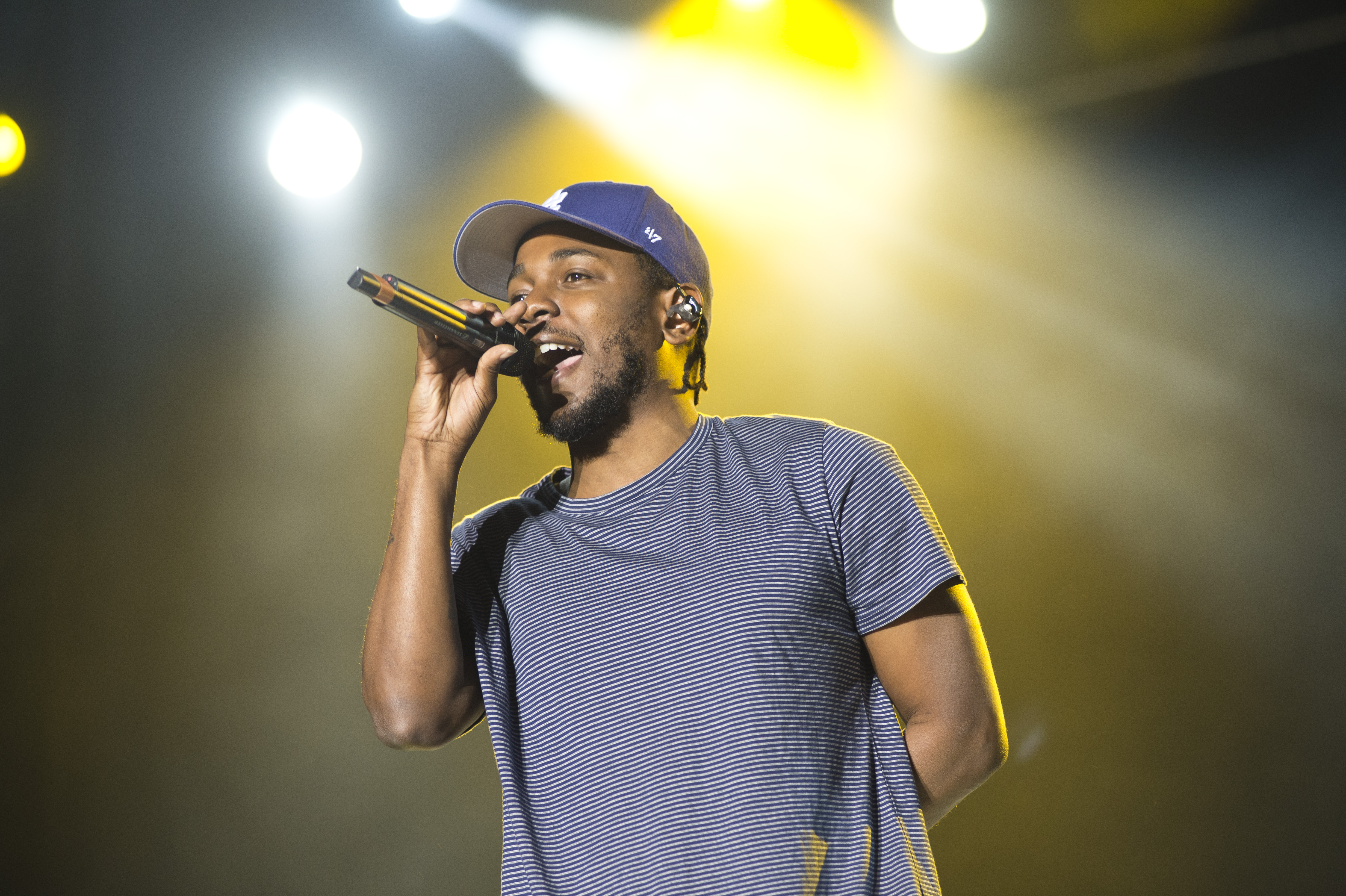 Kendrick Lamar performing on stage, wearing a striped shirt and blue cap, holding a microphone with bright stage lights behind him.