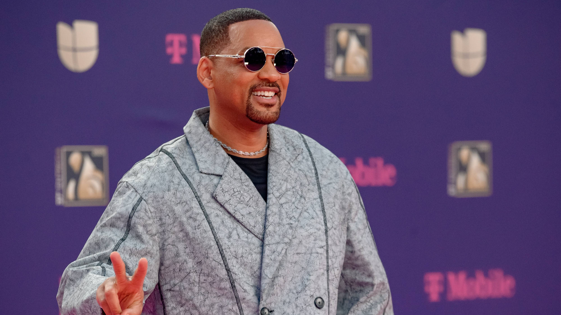 A man wearing sunglasses and a patterned coat, smiling and making a peace sign on a red carpet with a purple background.