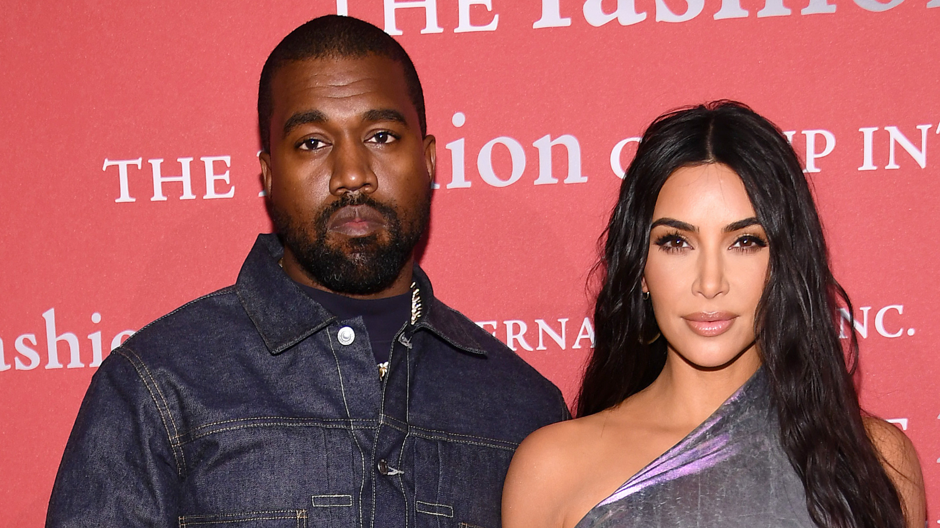 Kanye West and Kim Kardashian pose together at an event, standing in front of a red backdrop with text.