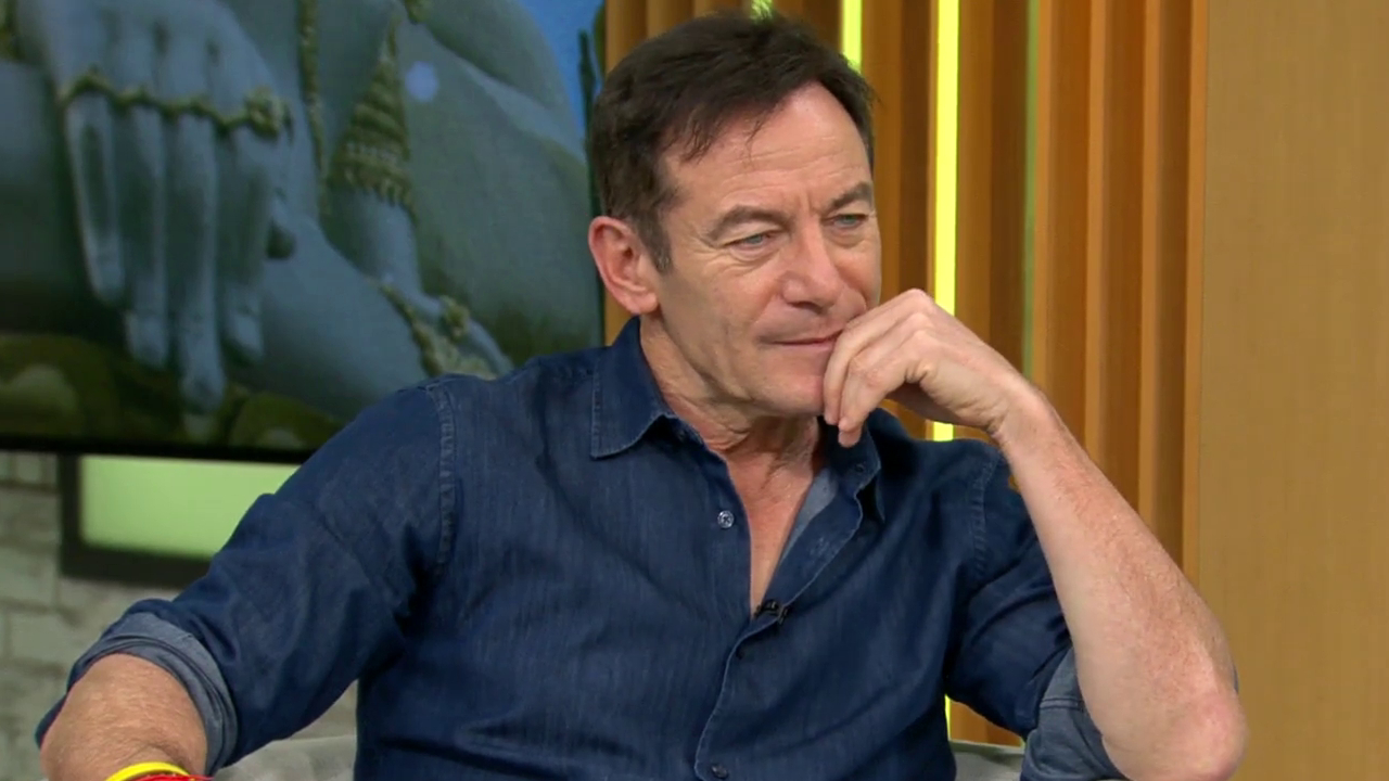 Jason Isaacs in a denim shirt sitting thoughtfully, with a hand on his chin, in a brightly lit room with a large image in the background.