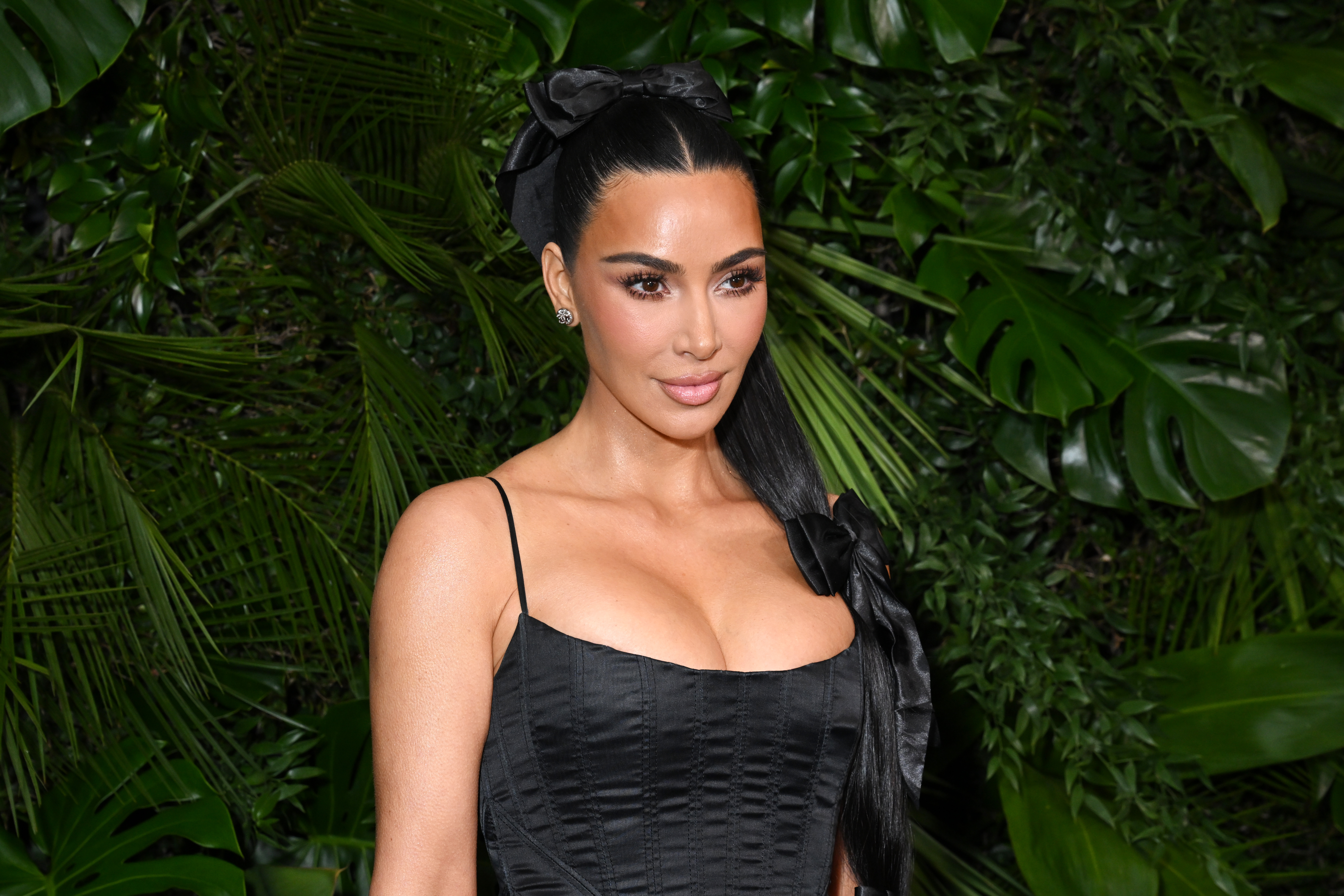 Kim Kardashian in a black dress with a bow in her hair, standing in front of lush green foliage.