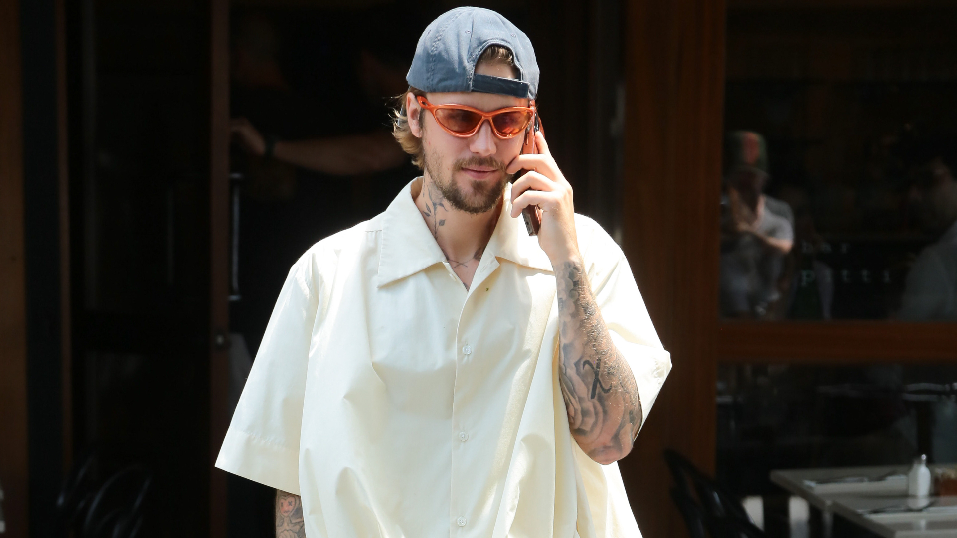 A man with tattoos, wearing a white shirt, orange sunglasses, and a backward cap, talks on the phone outside a building.