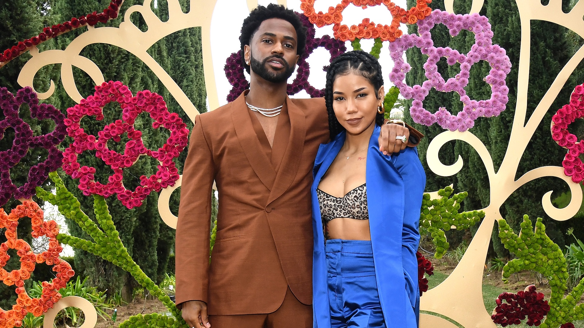 Big Sean in a brown suit and Jhené Aiko in a blue outfit stand together, posing in front of a floral backdrop.