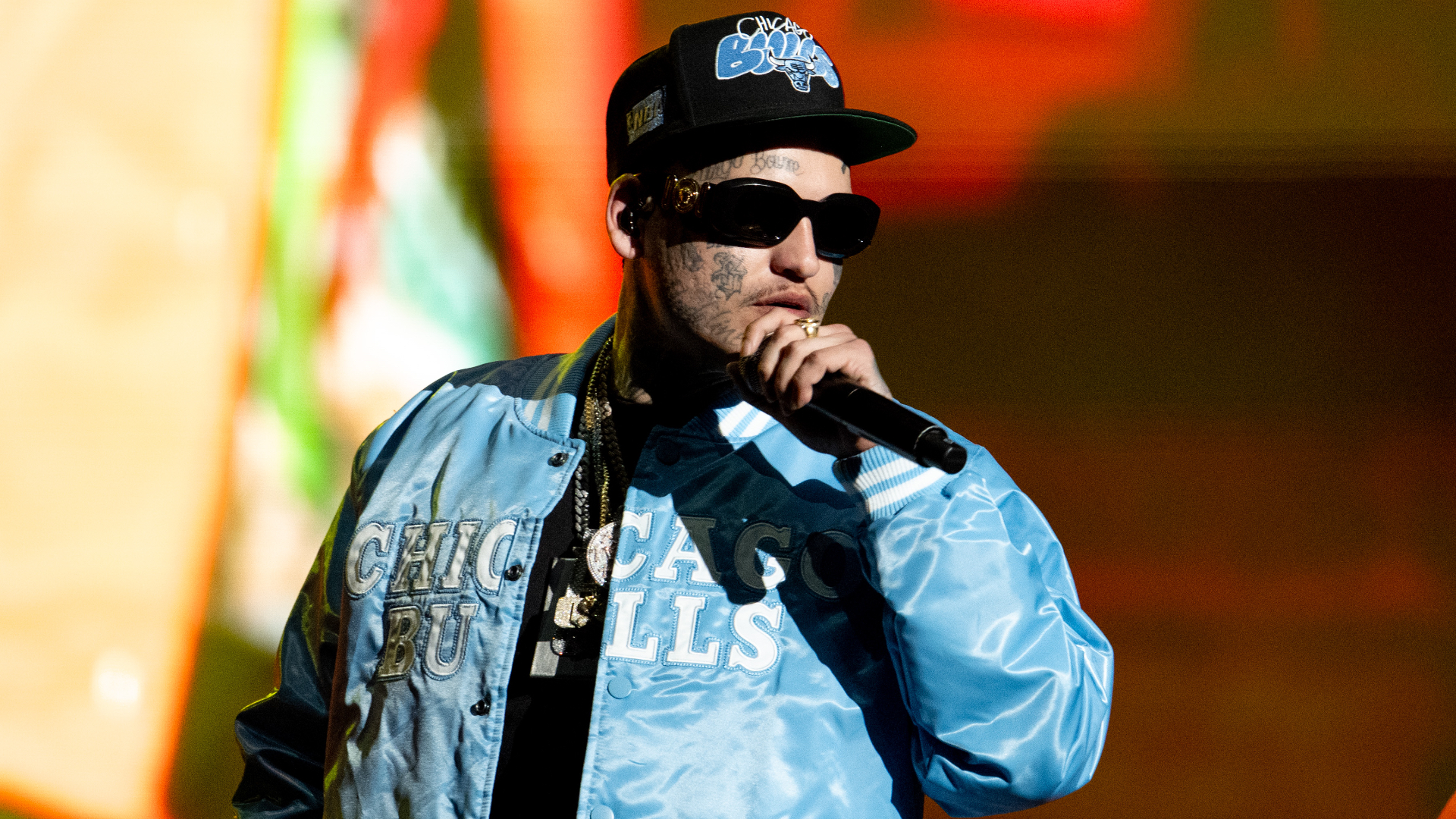 Lefty Gunplay wearing sunglasses, a Chicago Bulls jacket, and a cap, holding a microphone on stage.
