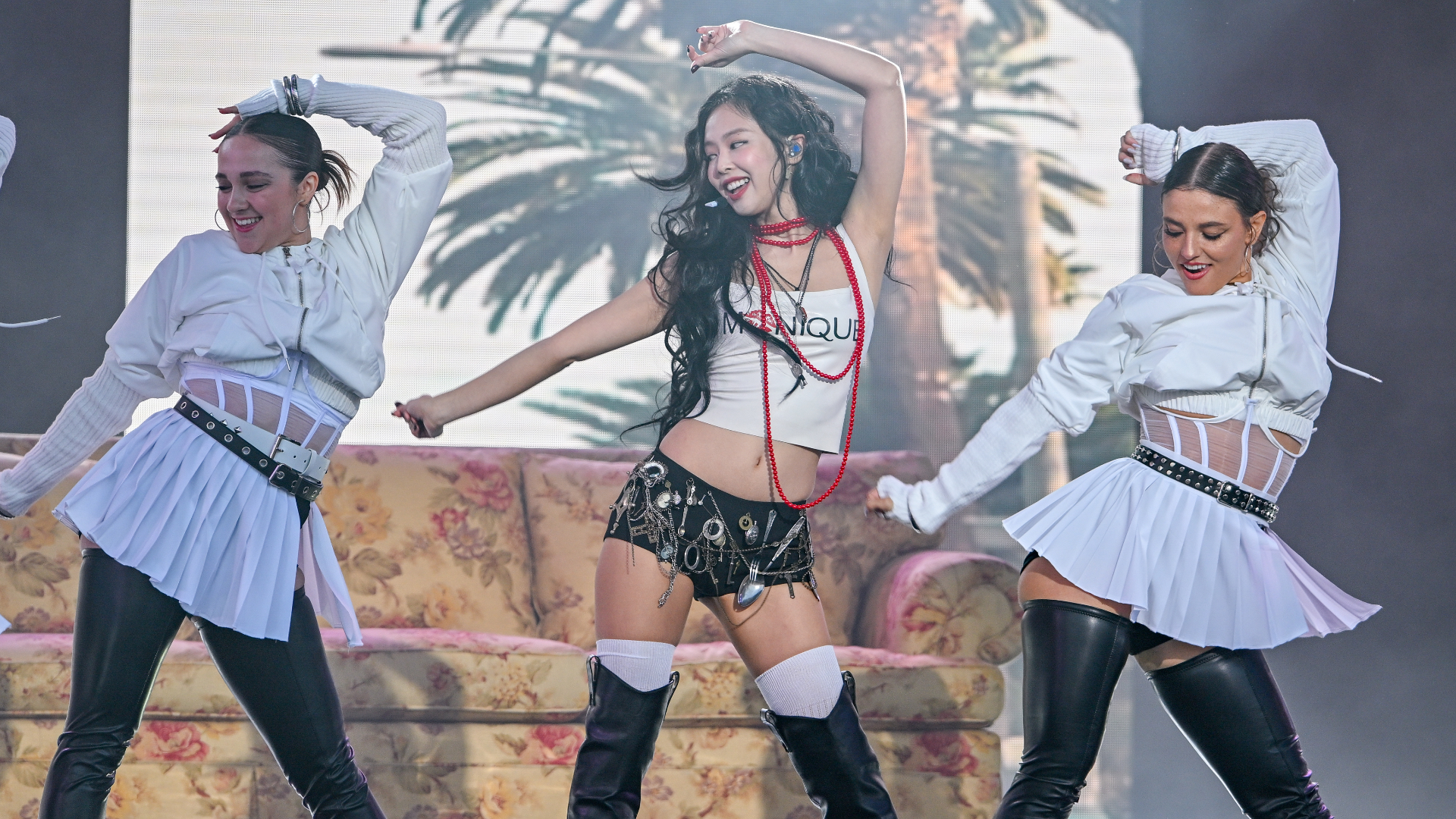 Jennie in a stylish outfit dances energetically with two backup dancers on stage, with a tropical backdrop.