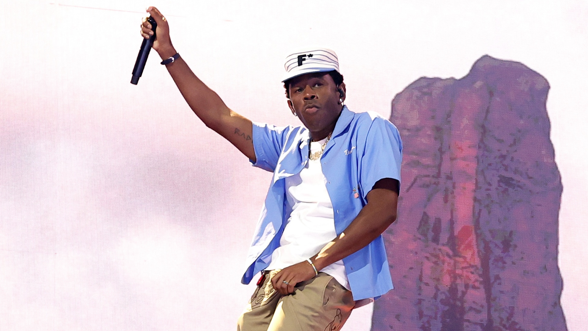 Tyler, the Creator performing on stage, wearing a blue shirt and white hat, holding a microphone with a scenic backdrop.