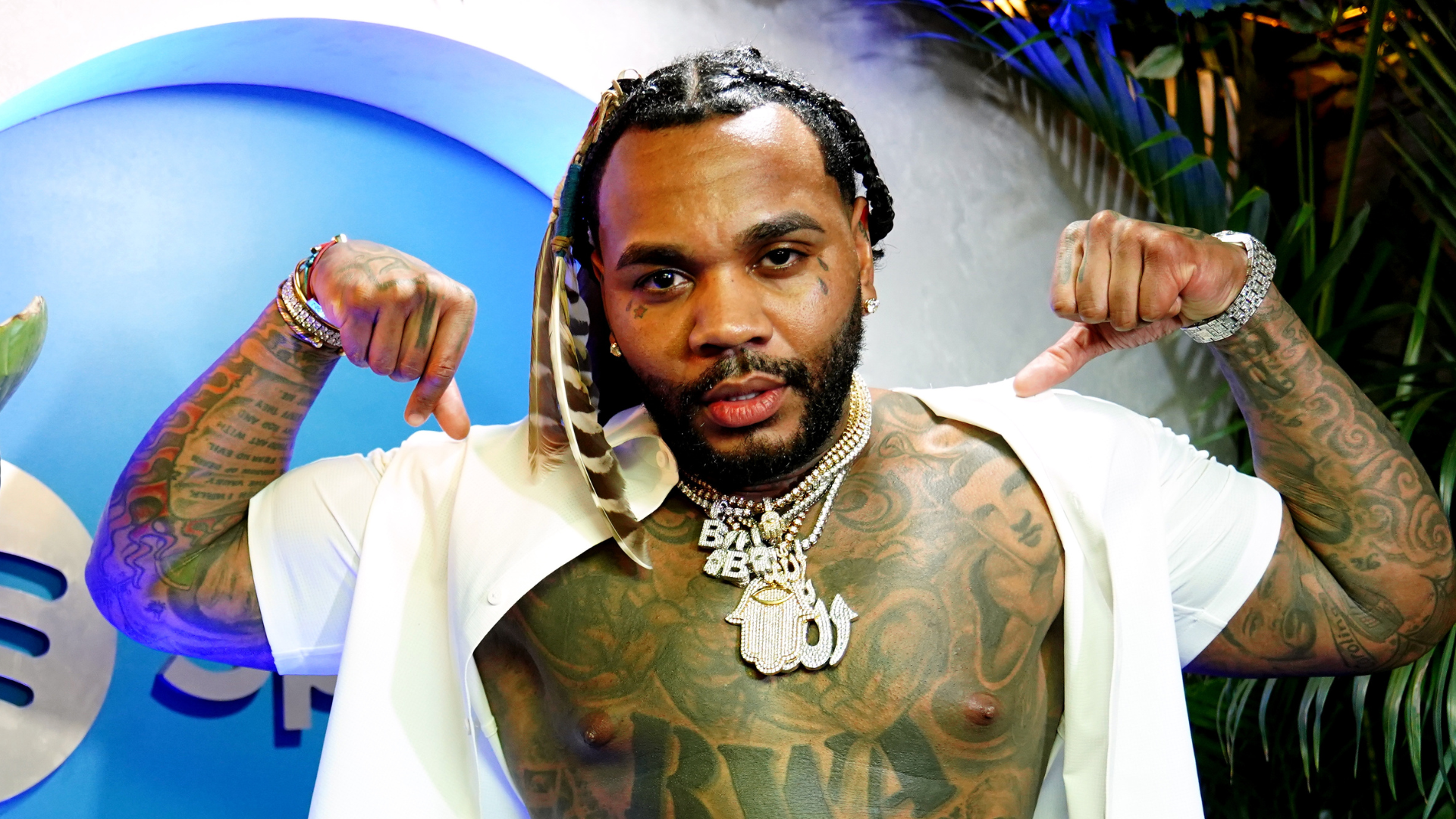 Kevin Gates flexing his muscles, shirt open, showcasing tattoos and jewelry, with a confident expression.