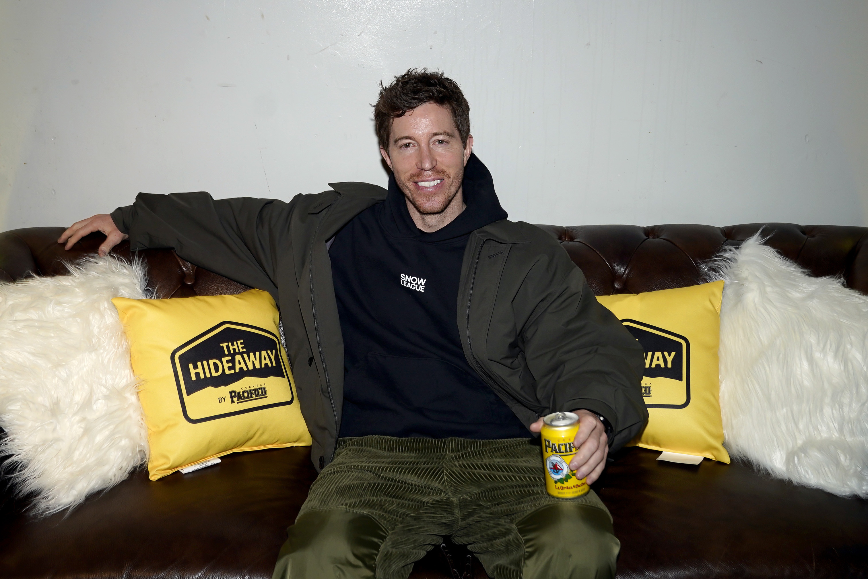 A man sitting on a couch with "The Hideaway" pillows, holding a can of Pacifico beer, wearing a dark hoodie and olive pants.