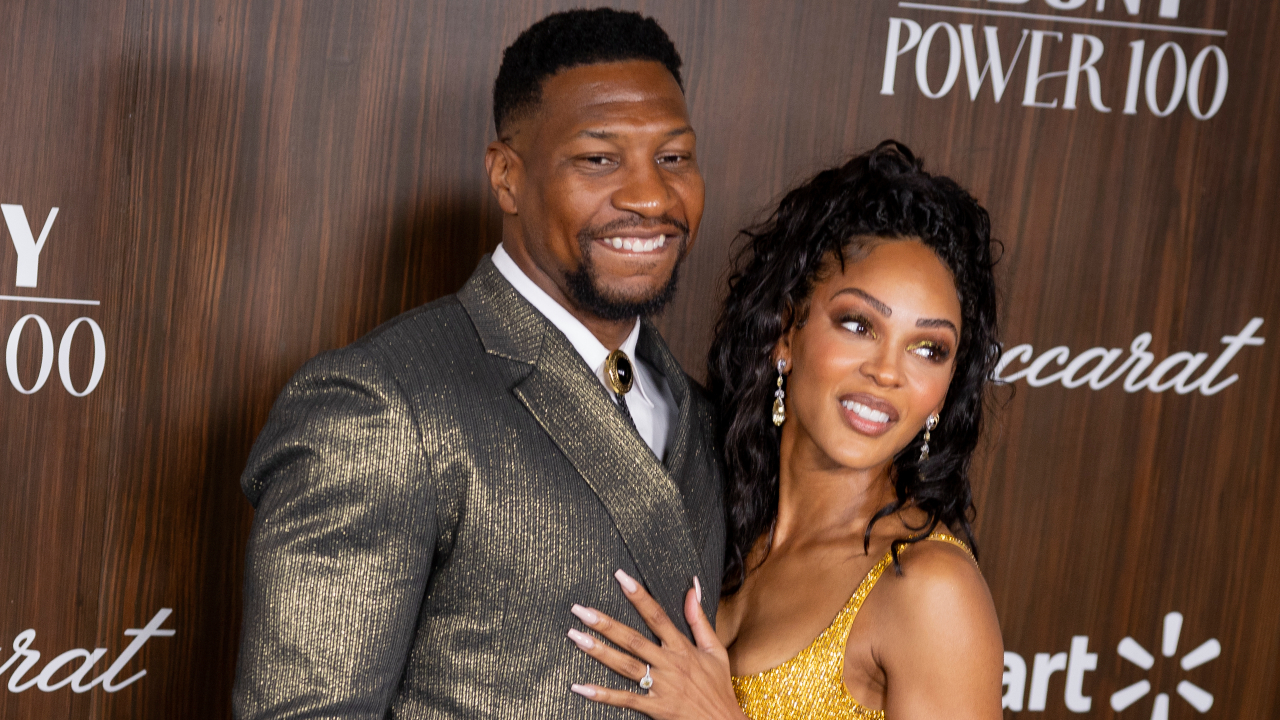 Jonathan Majors in a glittering suit and Meagan Good in a gold dress pose together, smiling at an event with a wooden backdrop.