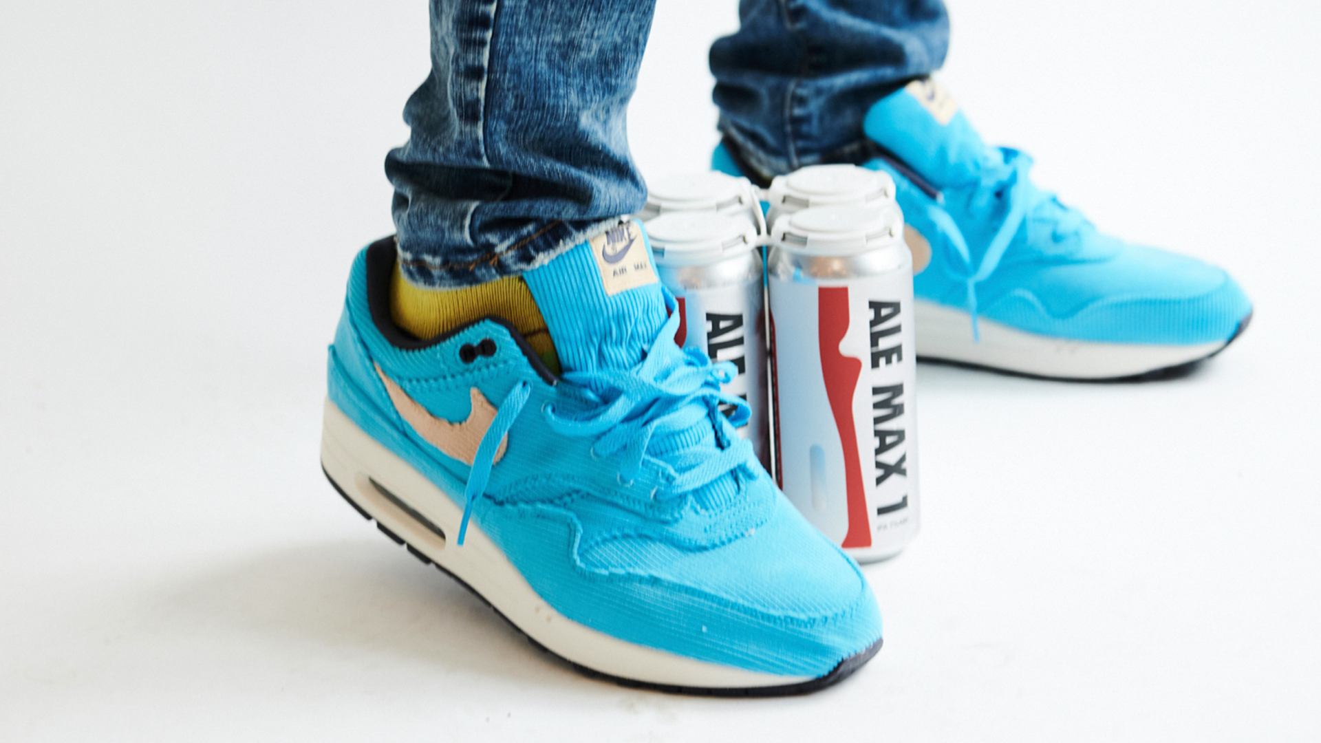 Blue sneakers with a pink swoosh, worn with jeans, next to cans labeled "Ale Max 1" on a white background.