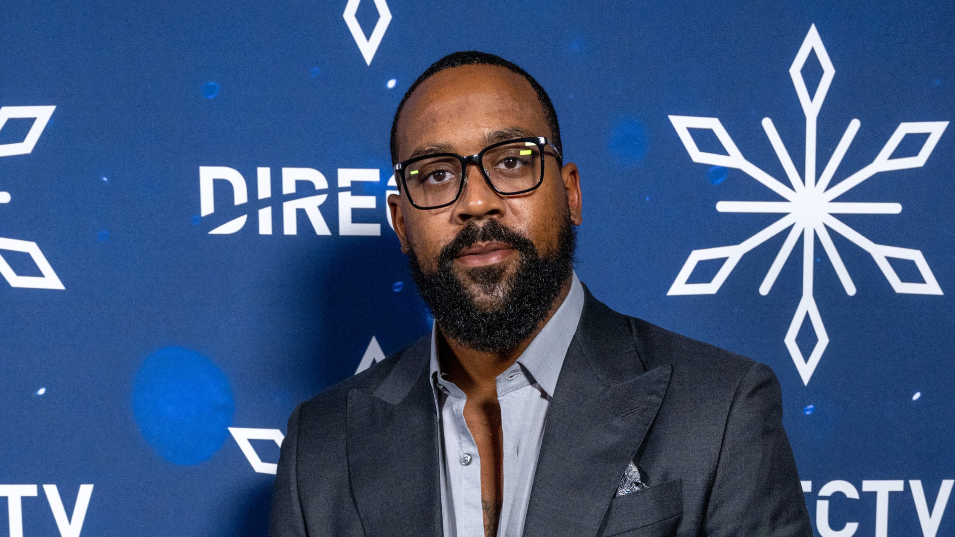 A man in a suit with glasses and a beard stands against a blue background with snowflake designs and the DirecTV logo.