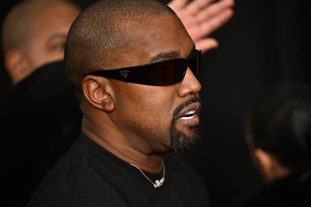 Kanye West wearing dark sunglasses and a black shirt, with a profile view against a dark background.