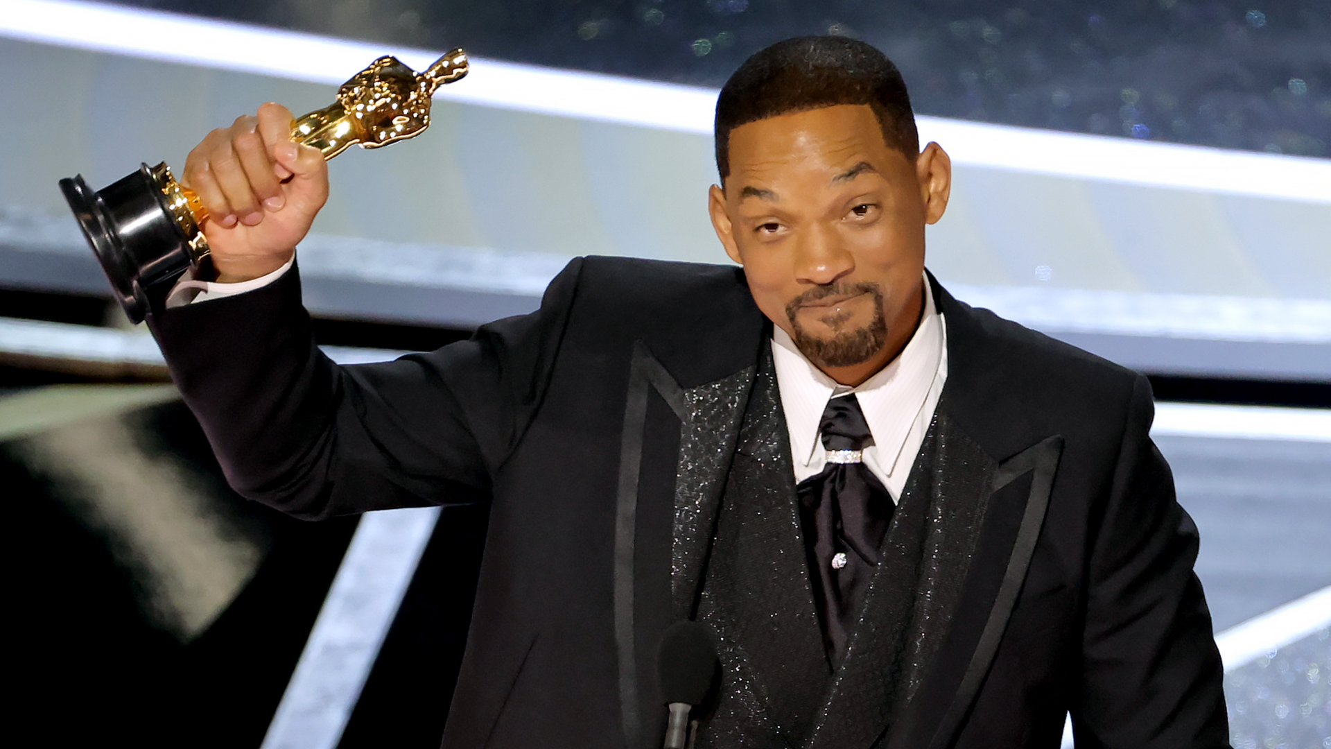 Will Smith holding an Oscar, wearing a black tuxedo, on stage at an award ceremony.