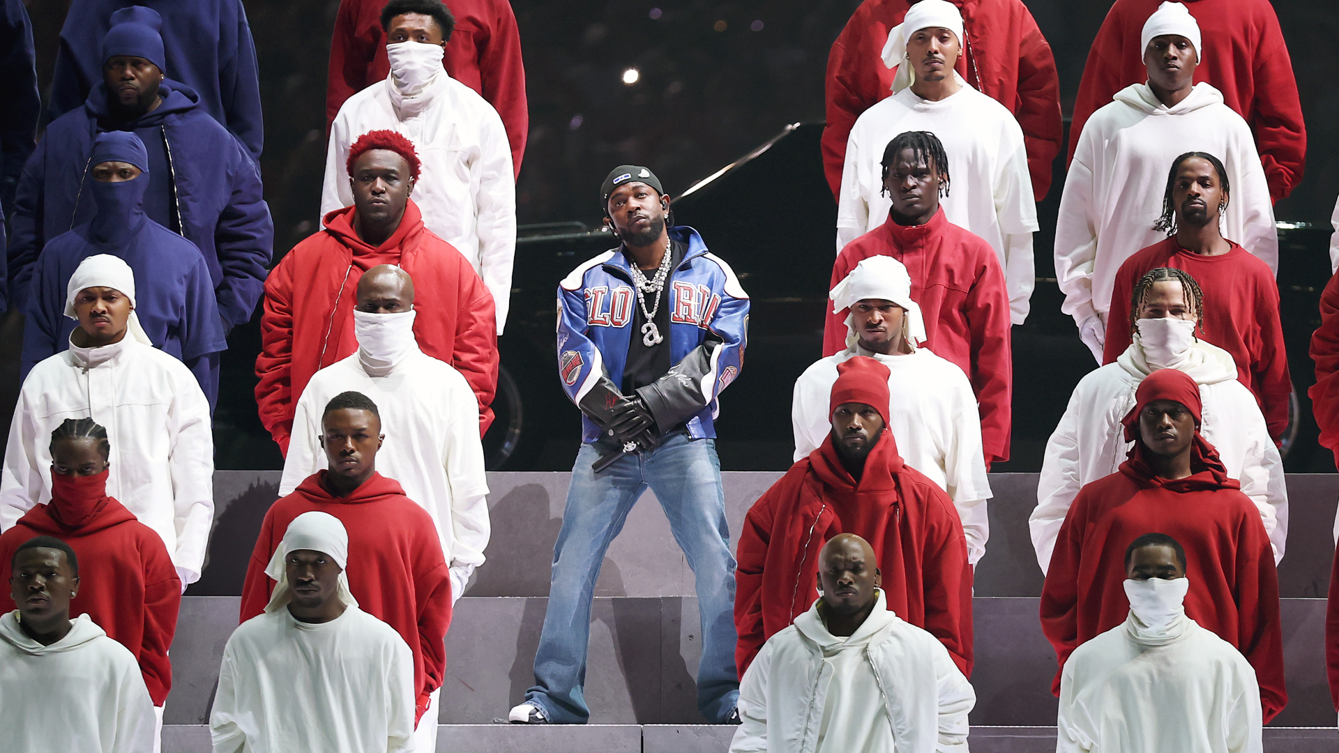 A group of performers in red, white, and blue outfits on a tiered stage. Central figure is Kendrick Lamar in a blue jacket stands out.