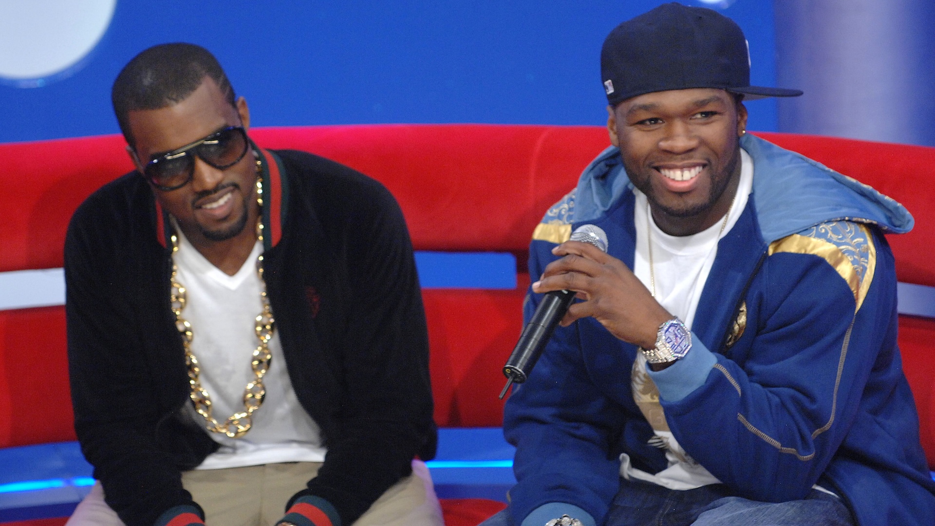 Kanye West and 50 Cent sitting on a red couch, smiling. Kanye wears sunglasses and a chain, while 50 Cent holds a microphone.