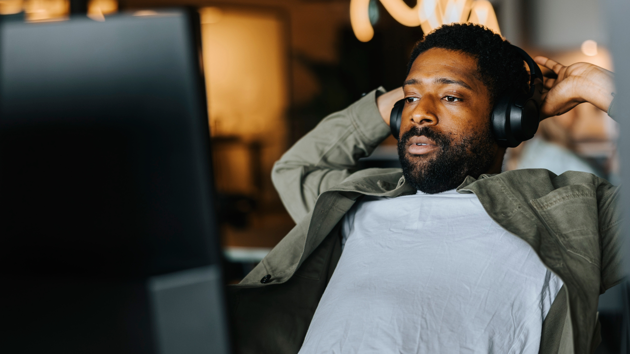 A man wearing headphones, sitting back and looking at a computer screen, appears stressed in a modern office setting.