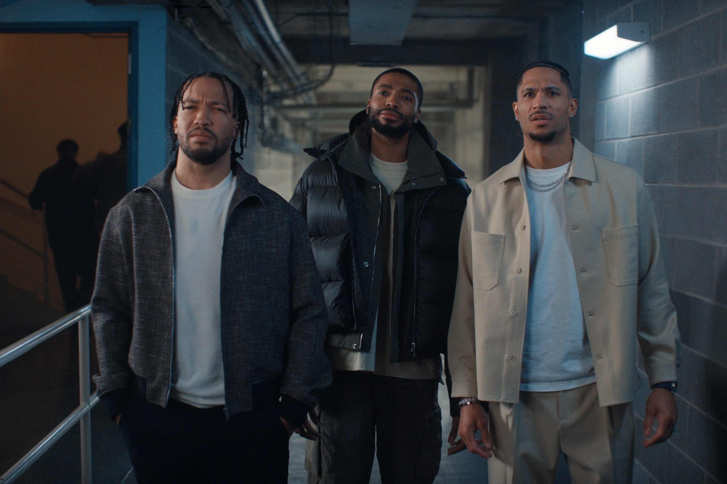 Josh Hart, Jalen Bruson, and Mikal Bridges stand in a dimly lit hallway, dressed in casual and stylish jackets, looking intently forward.