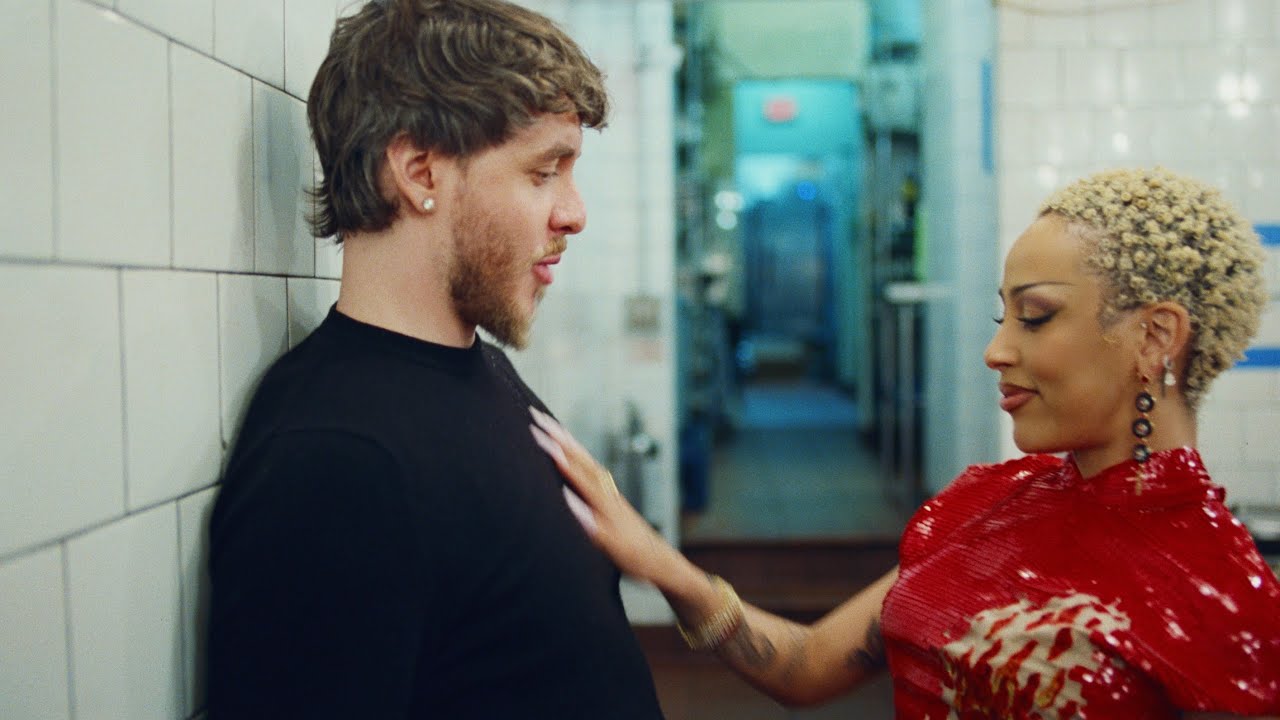 Jack Harlow in a black shirt leans against a tiled wall while Doja Cat, in a red outfit with short curly hair, touches his chest. They appear to be in a kitchen.