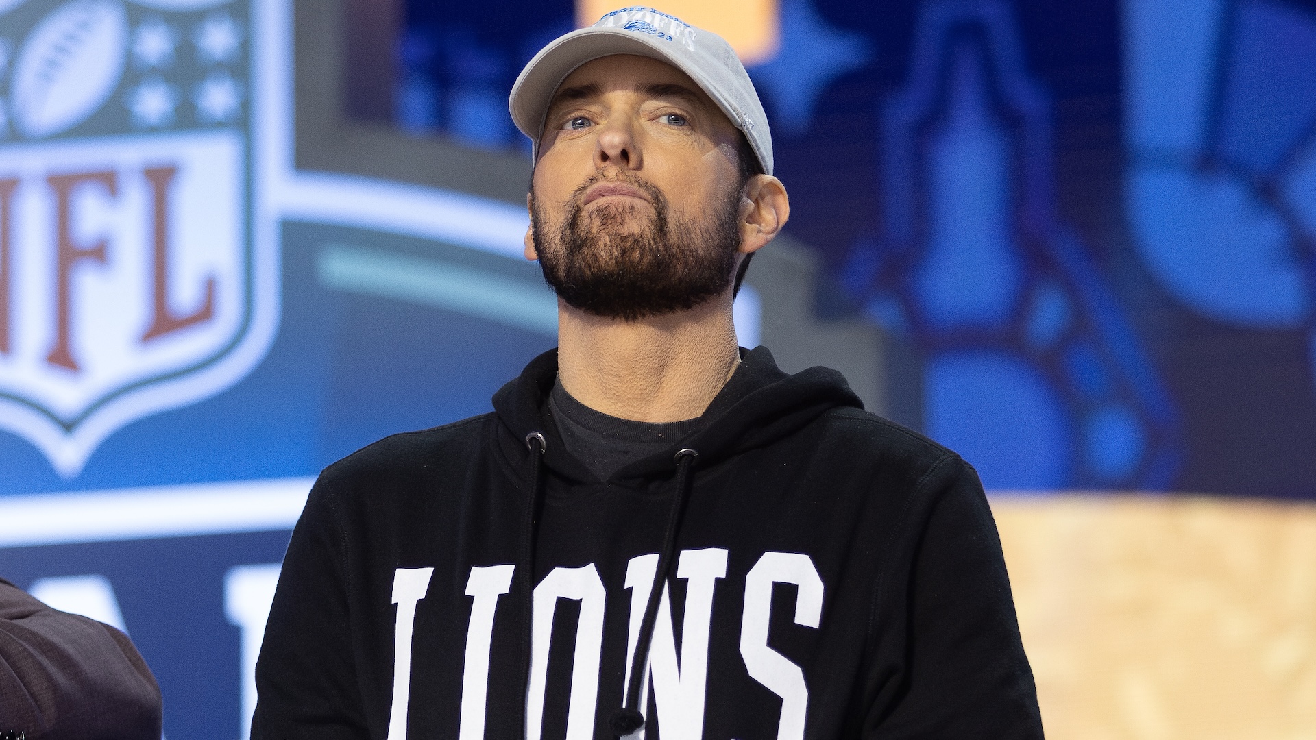 Eminem wearing a "Lions" hoodie and cap, standing in front of an NFL-themed background.