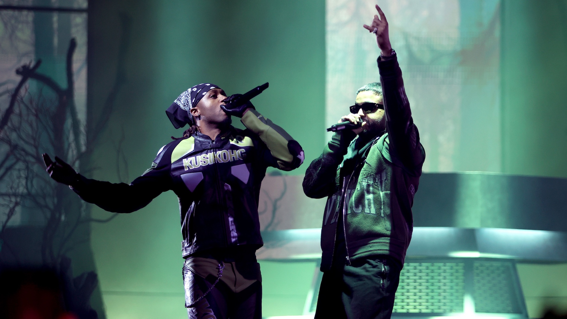 Two performers on stage, one wearing a bandana and jacket with "Kushok," the other in sunglasses and a jacket, both holding microphones.