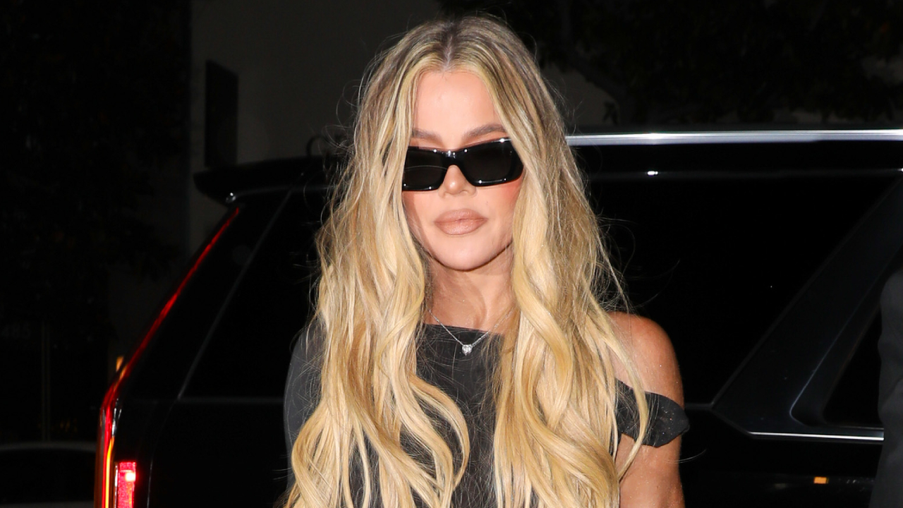 Khloé Kardashian with long blonde hair and sunglasses stands in front of a car at night, wearing a black outfit.
