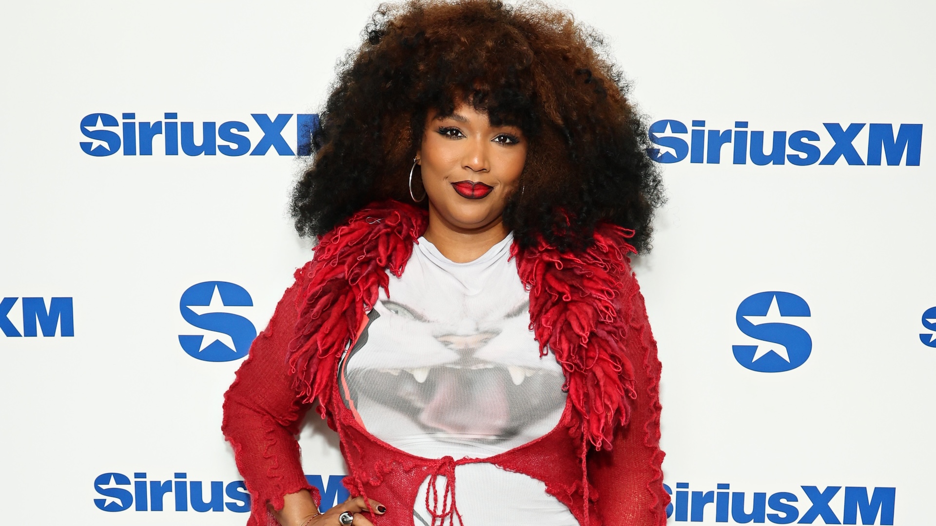 Lizzo at a SiriusXM event, wearing a red jacket and white shirt with bold makeup and curly hair.
