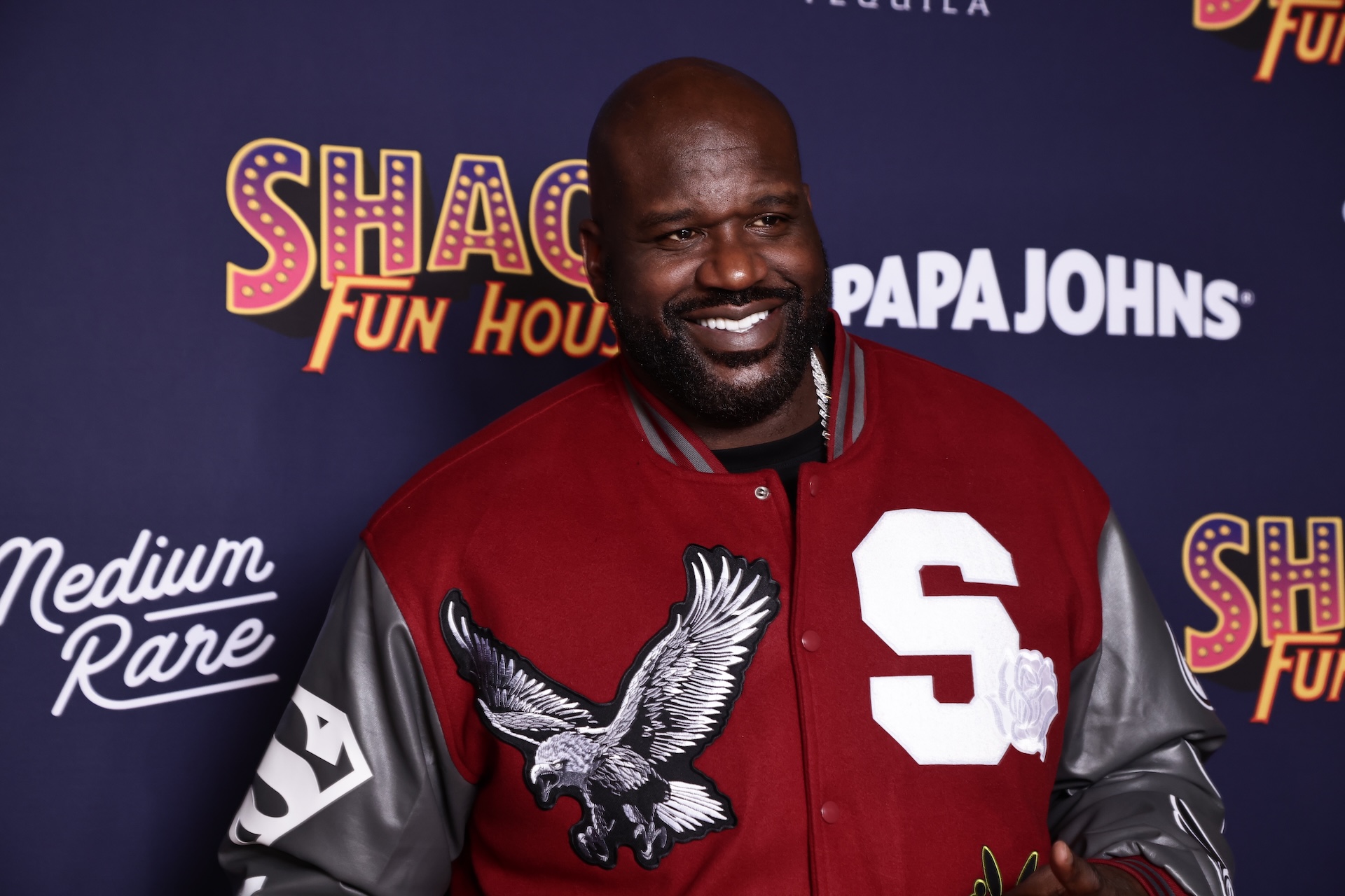Shaquille O'Neal smiling at an event, wearing a red varsity jacket with an eagle design, standing in front of branded signage.