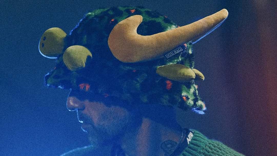 A person wearing a unique, horned hat with a green and yellow plush design, partially obscured in dim lighting. Nike x Cactus Plant Flea Market