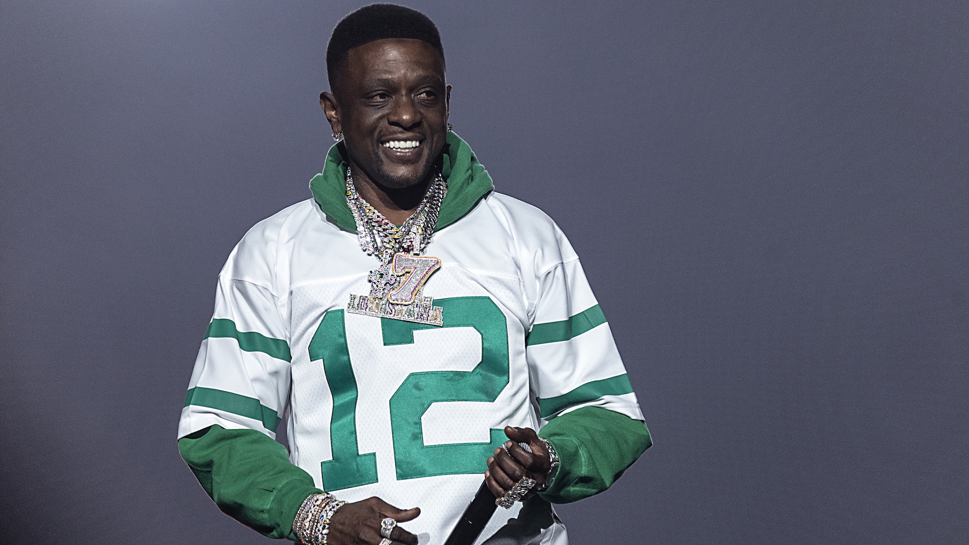 Rapper Boosie Badazz wearing a white and green sports jersey with the number 12, adorned with multiple chains, smiling on stage.