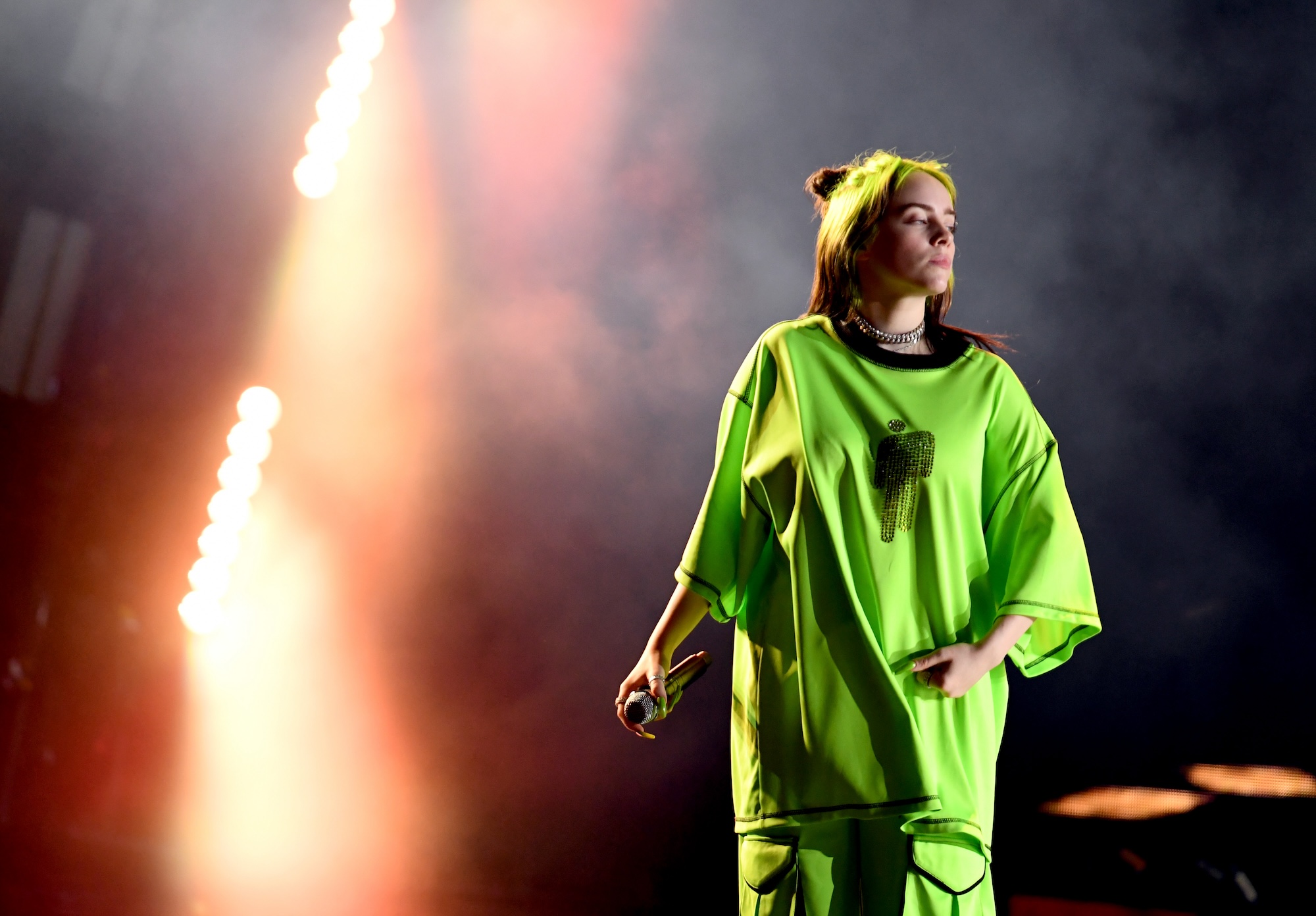 Billie Eilish performing on stage, wearing a neon green outfit, with dramatic lighting and smoke in the background.