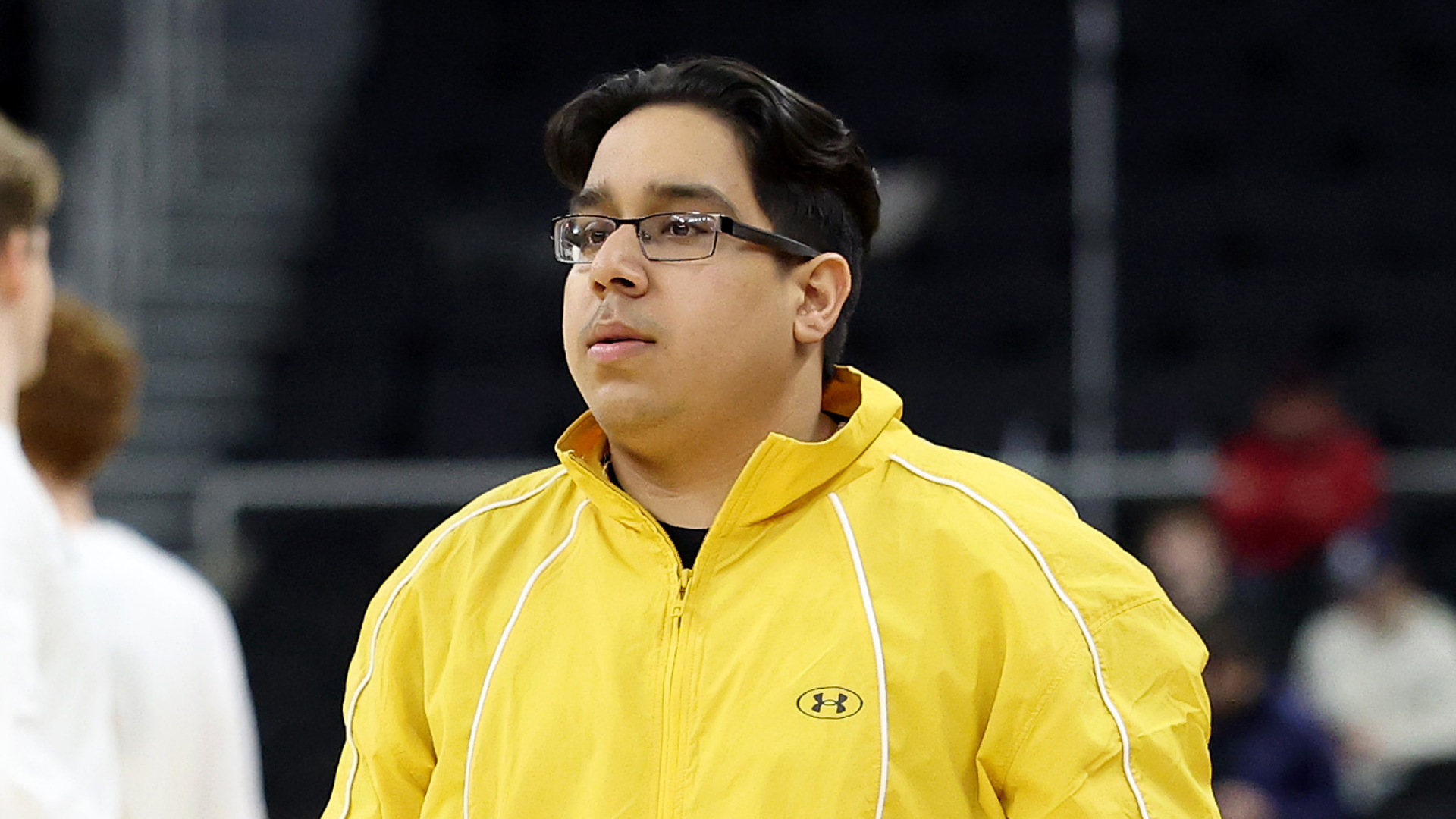 A person in a yellow jacket stands in a sports arena, looking to the side. The background is blurred, with people in the distance.