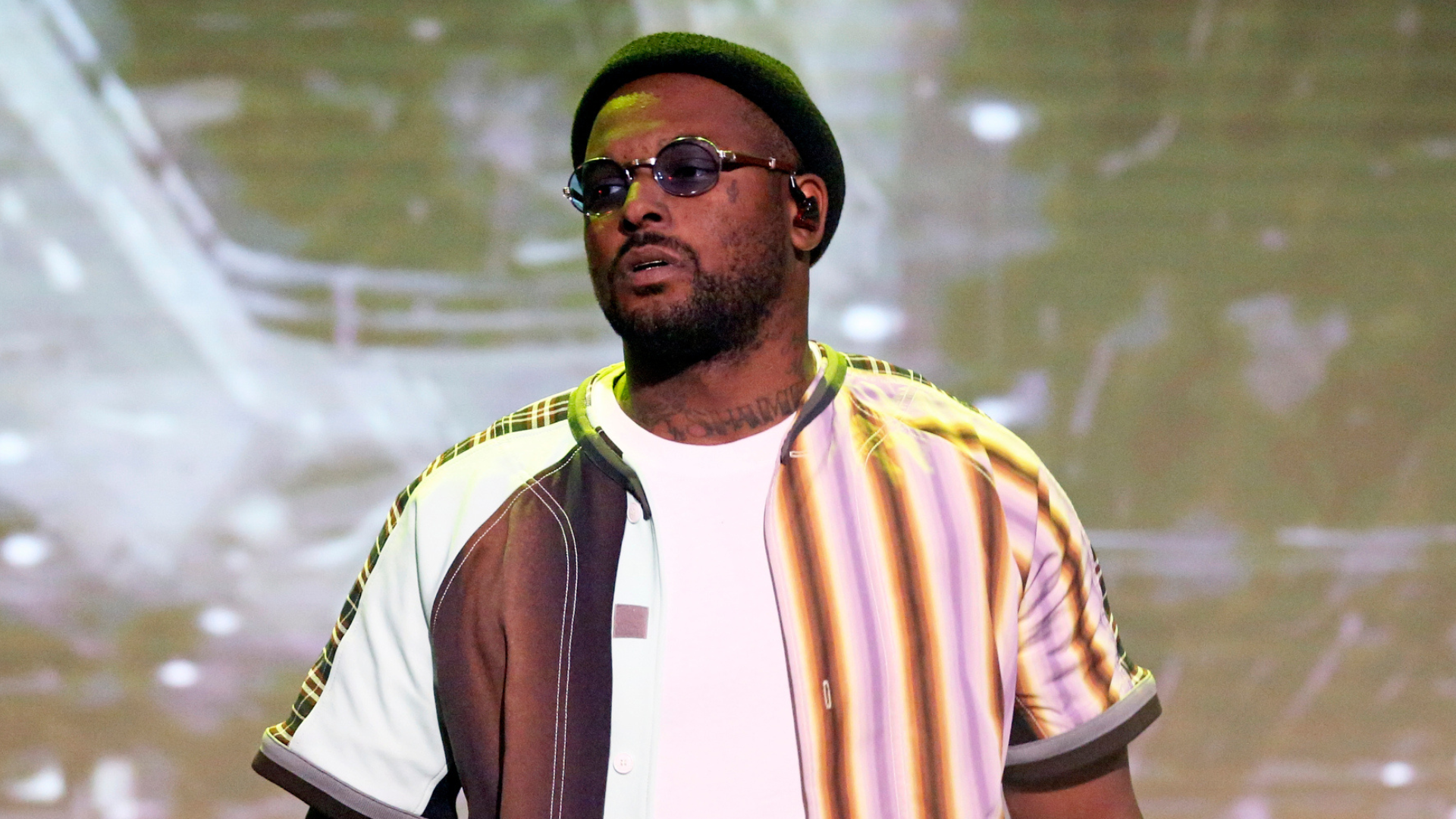 A man wearing sunglasses, a beanie, and a striped shirt stands against a blurred background.