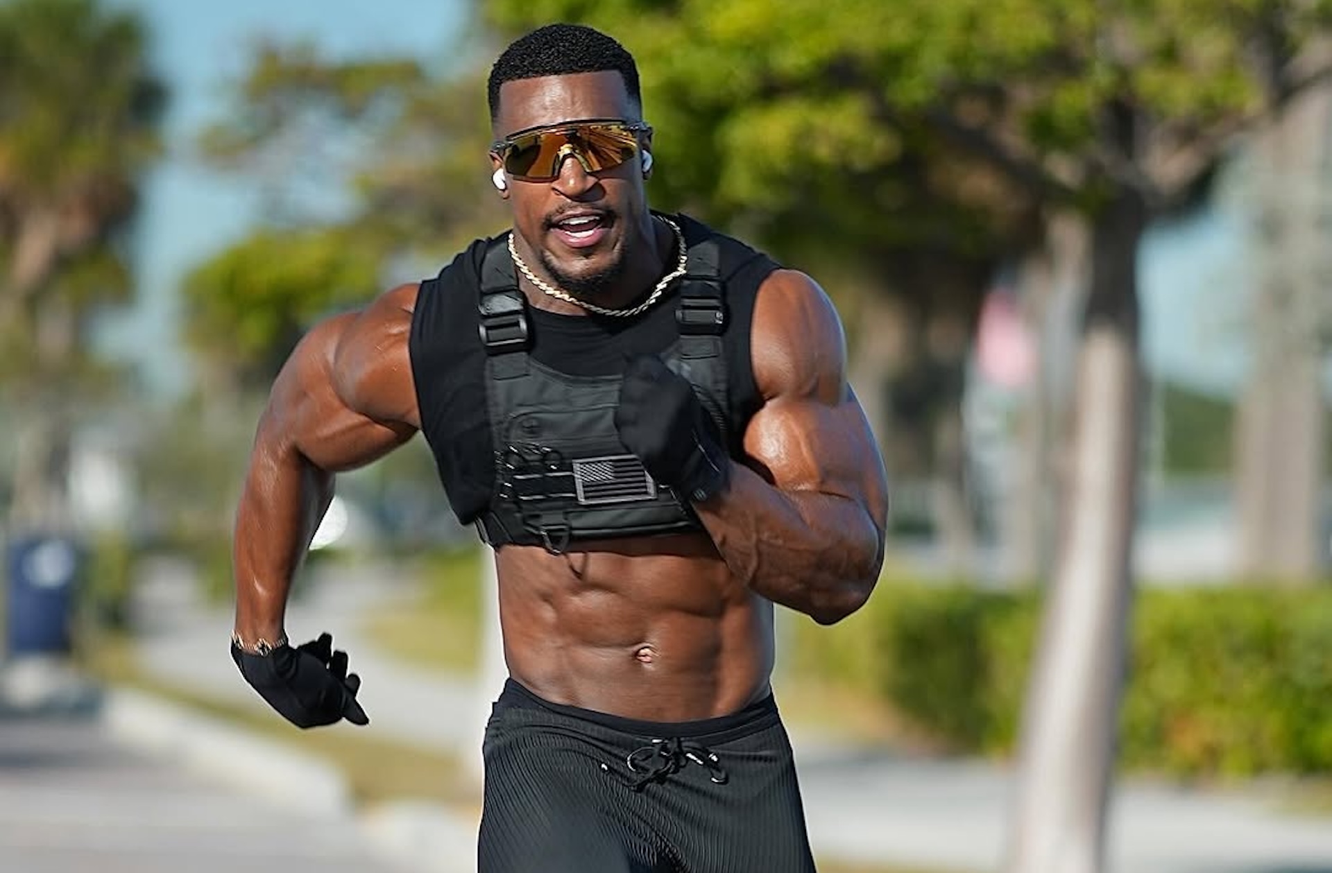 A muscular man running outdoors, wearing sunglasses and a weighted vest, with trees and a blurred background.