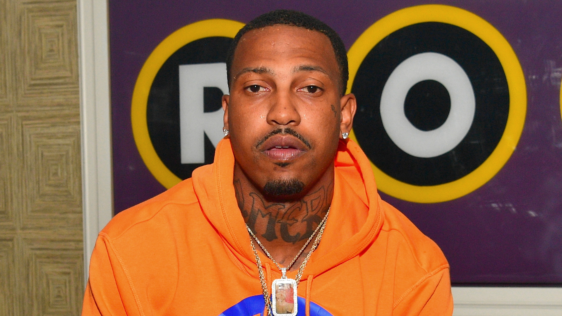 A man in an orange hoodie with tattoos and a chain necklace, standing in front of a sign with "RO" on it.