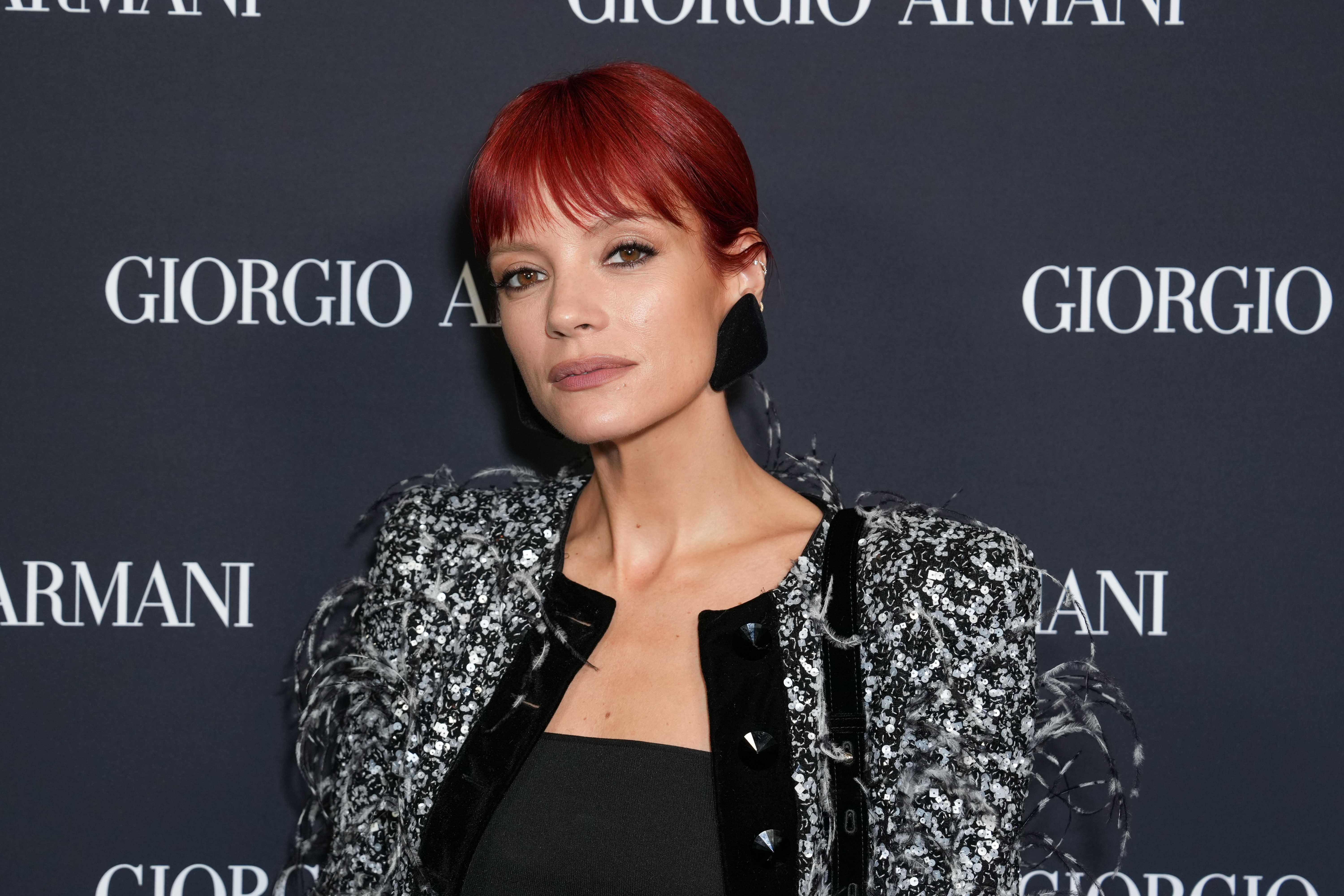 Lily Allen with red hair poses in front of a backdrop, wearing a textured black and silver outfit.