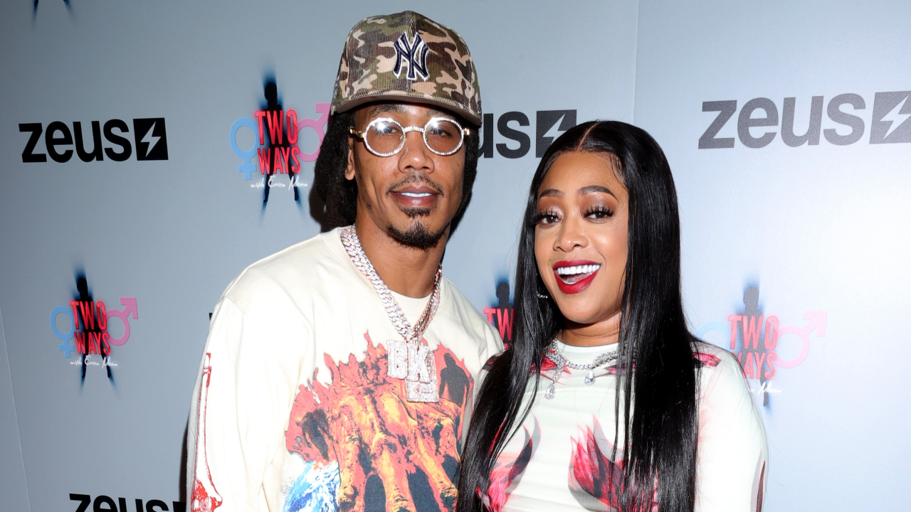Swurv in a cap and glasses stands next to wife Trina with long hair, both smiling at an event.