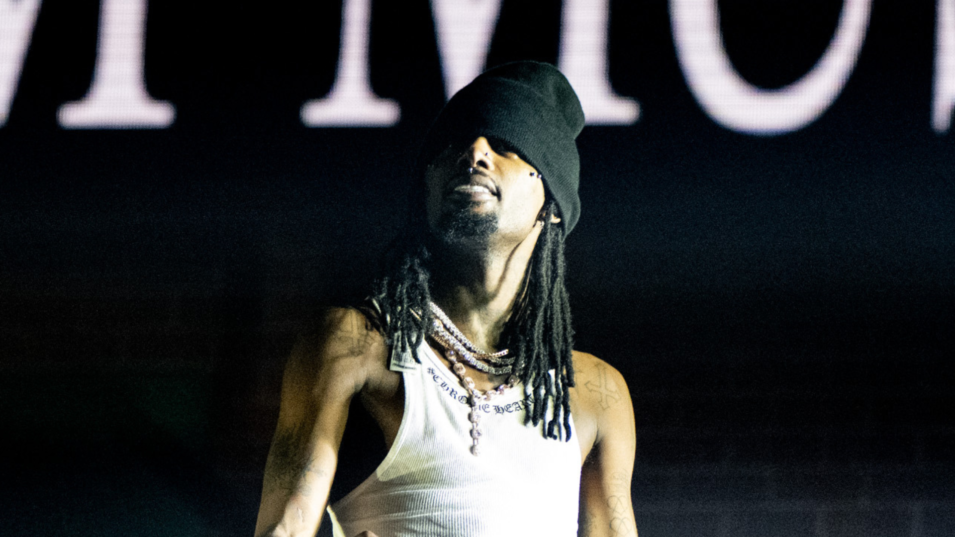 A man with long hair and a beanie performs on stage, wearing a white tank top and necklaces.