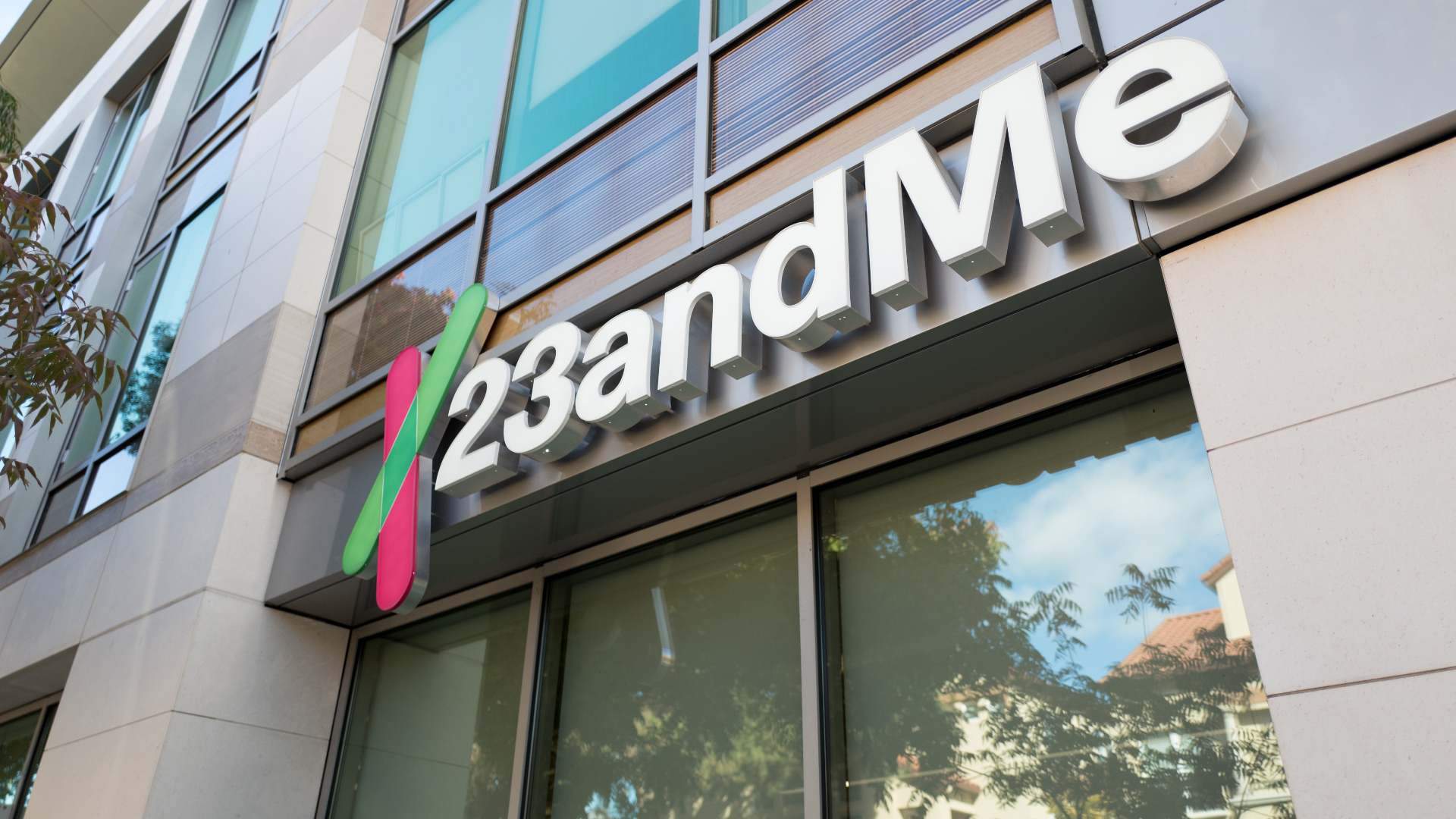 Sign of 23andMe on a modern building facade with large windows and reflections of trees and sky.