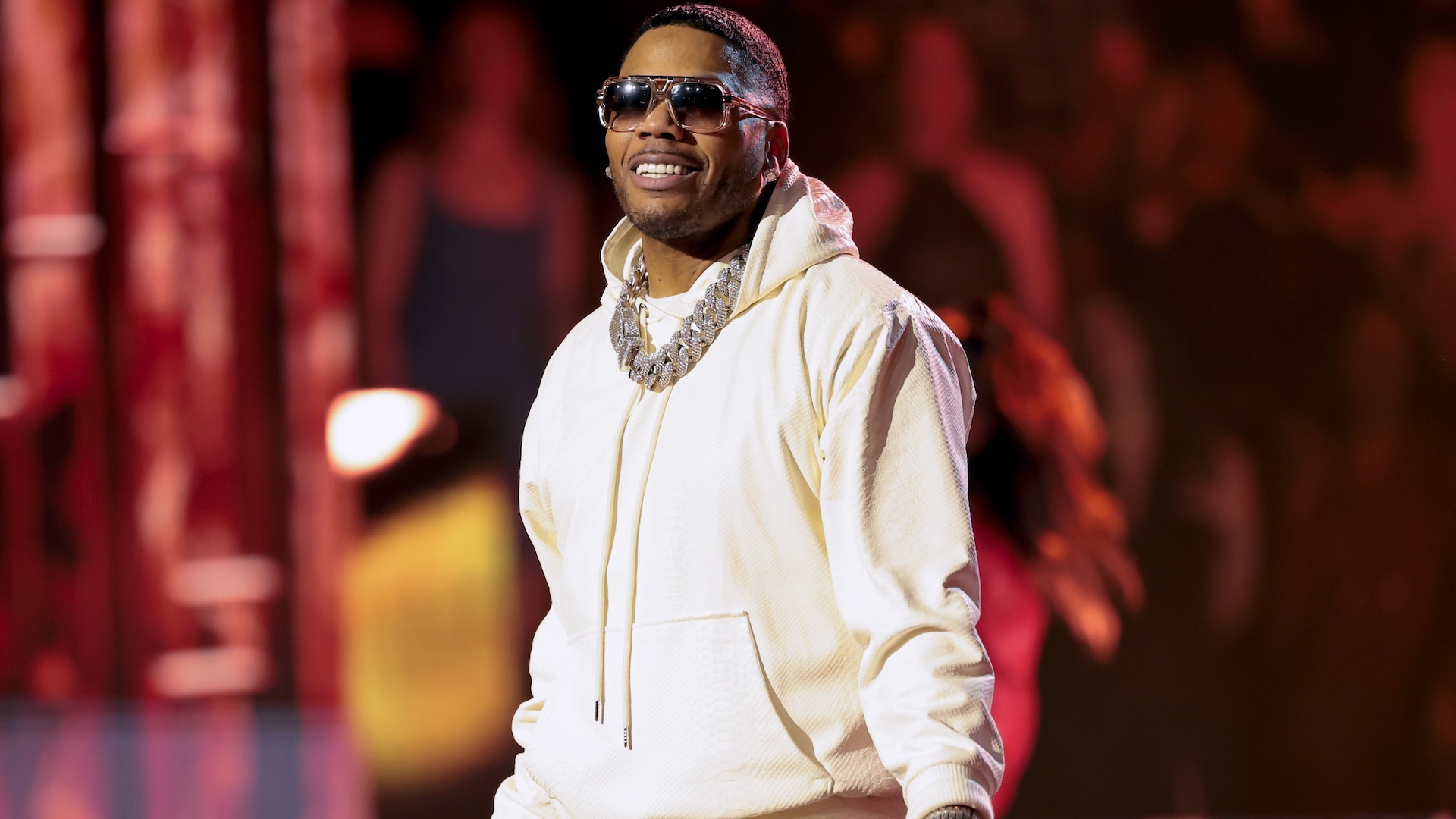 Rapper Nelly wearing sunglasses and a white hoodie, performing on stage with a vibrant background.