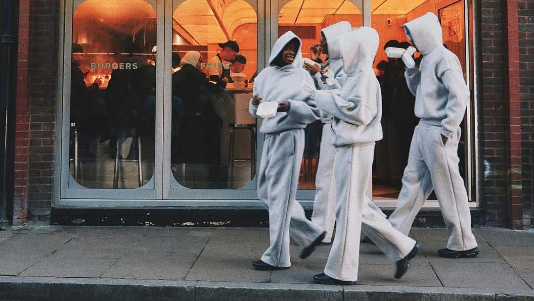 Four people in oversized gray hoodies walk past a restaurant, drinking from cups. The restaurant has large windows and people inside. Seventh