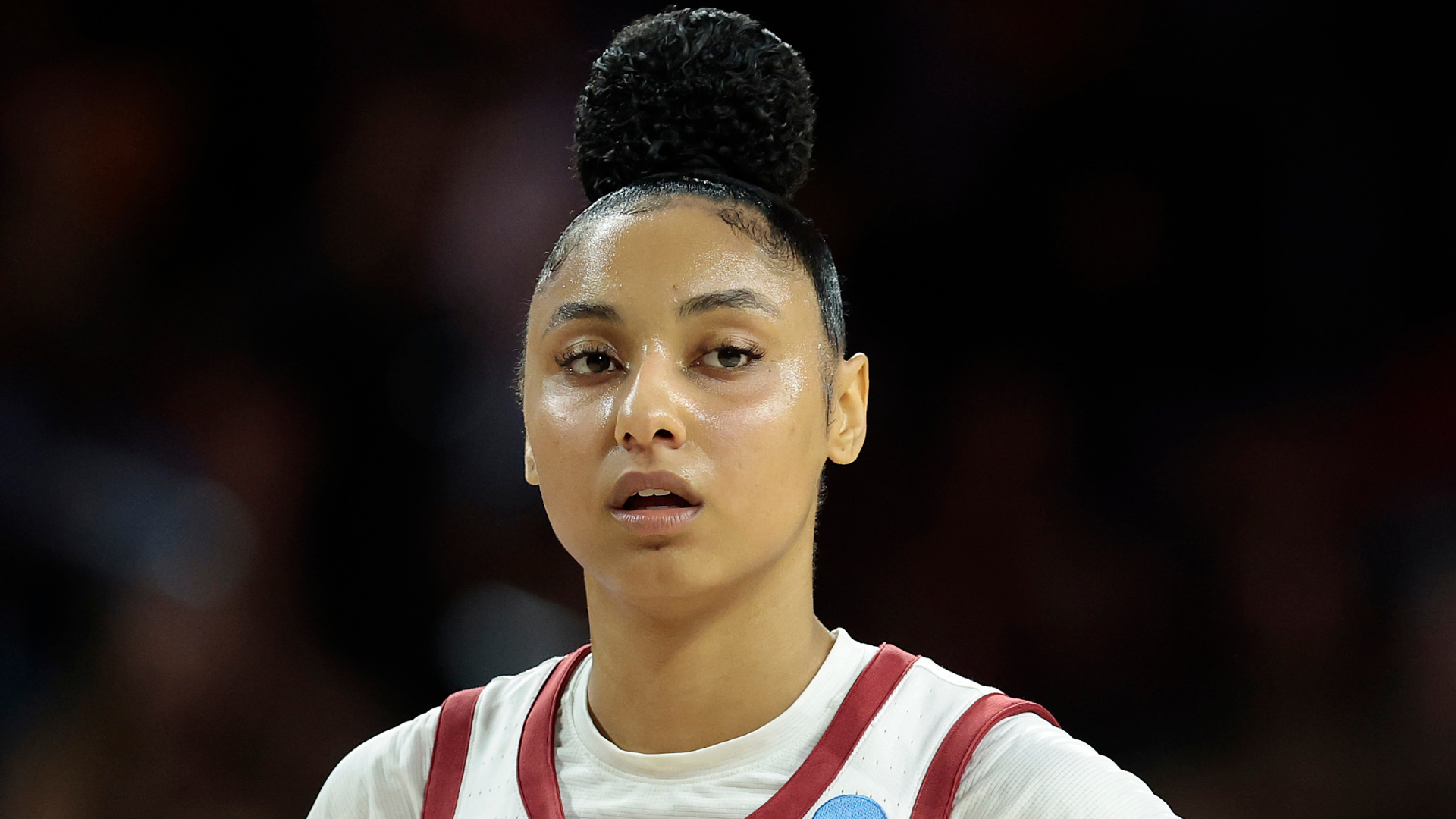 A female basketball player on the court, wearing a white jersey with red trim, hair in a bun, looking focused.