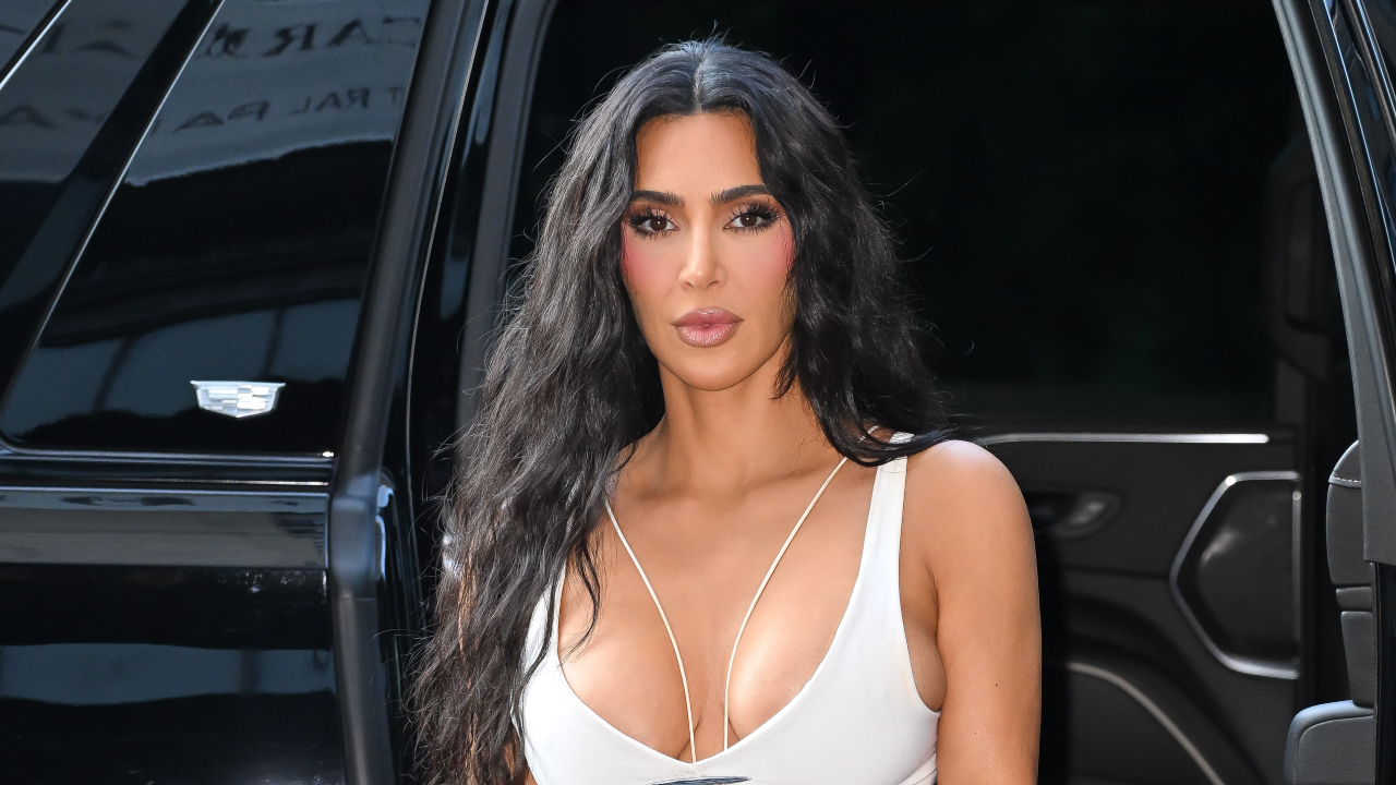 Kim Kardashian with long black hair and a white outfit, standing beside a black vehicle.