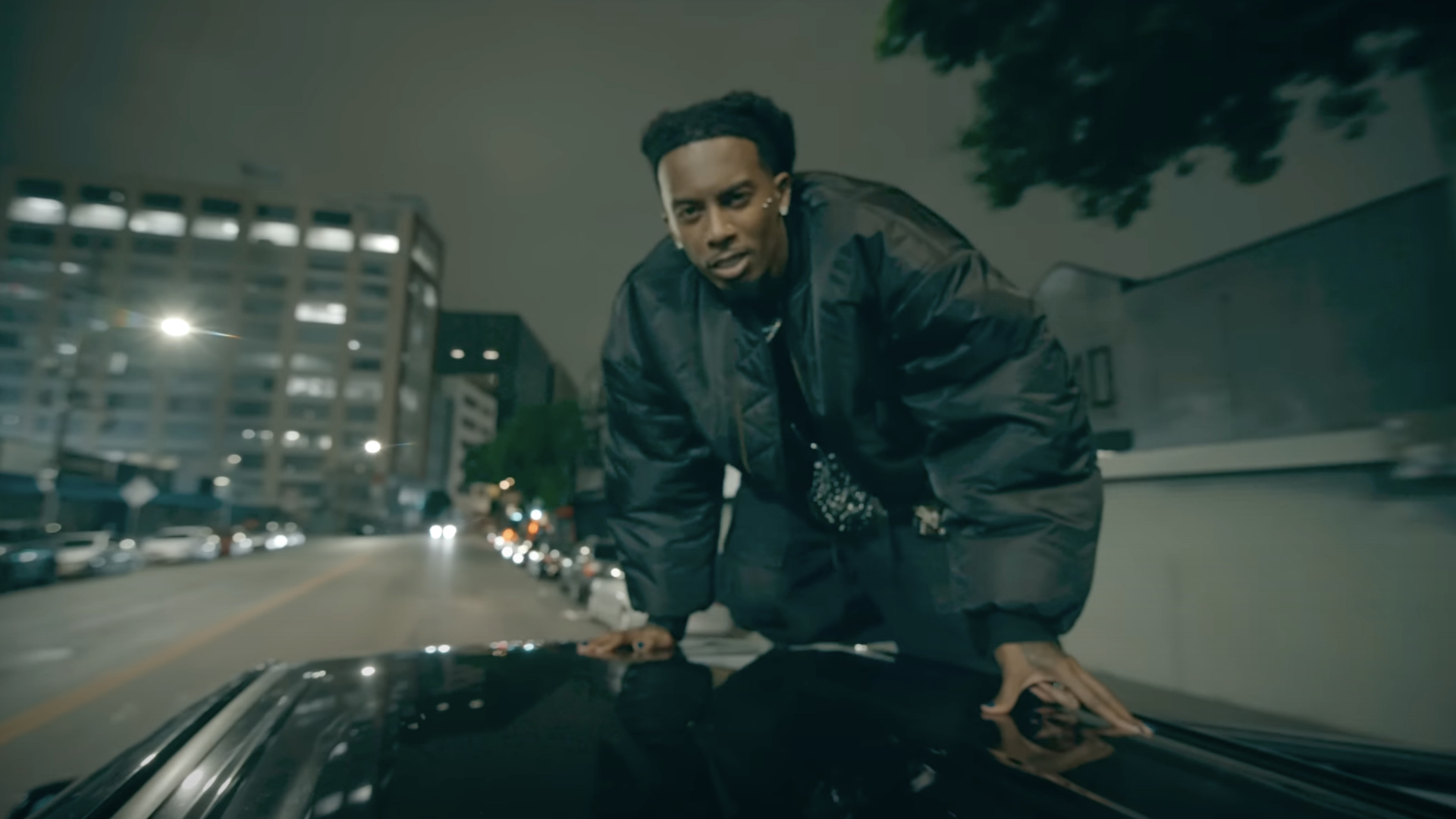 Playboi Carti in a dark jacket is crouched on the roof of a car at night, with city lights and buildings in the background.