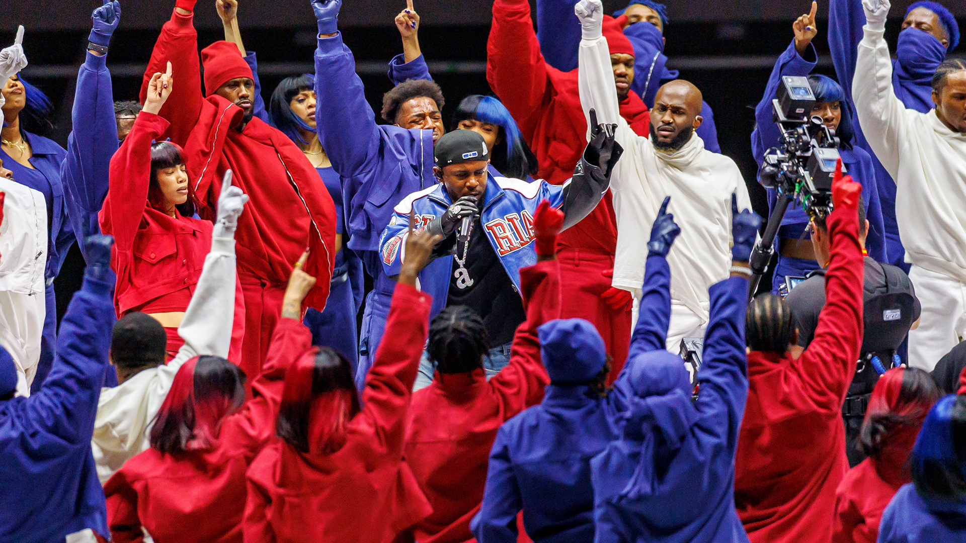 A group of performers in red, white, and blue outfits, energetically raising their hands. A central figure with a microphone, Kendrick Lamar, leads them.