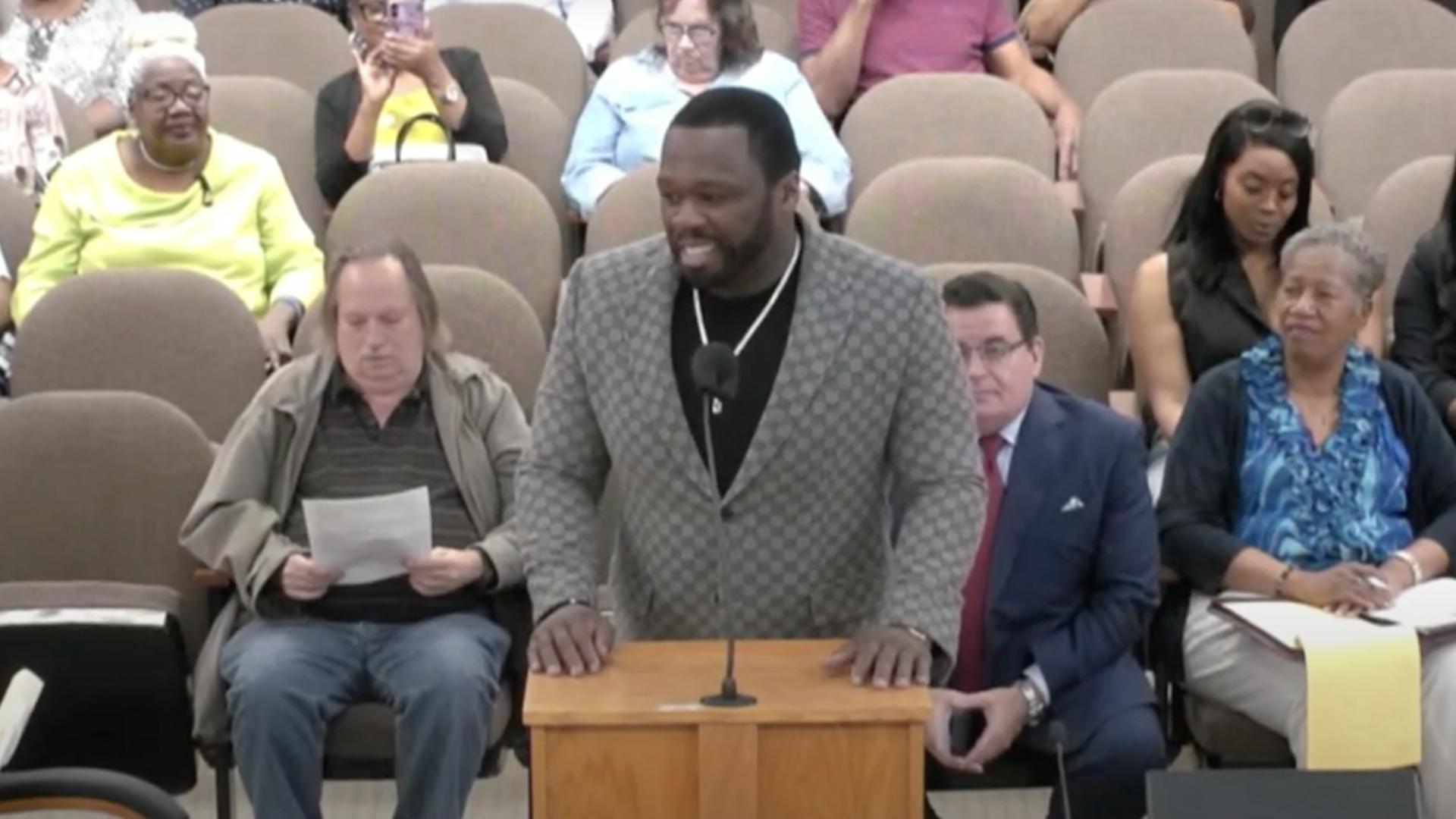 50 Cent speaks at a podium in a meeting room with an audience seated behind him.