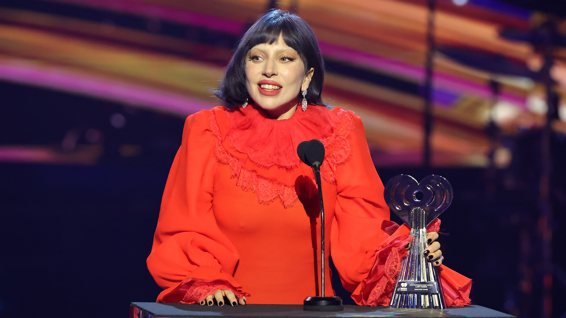 Lady Gaga in a red dress with a ruffled collar, holding a trophy, speaking at an event with colorful lights in the background.