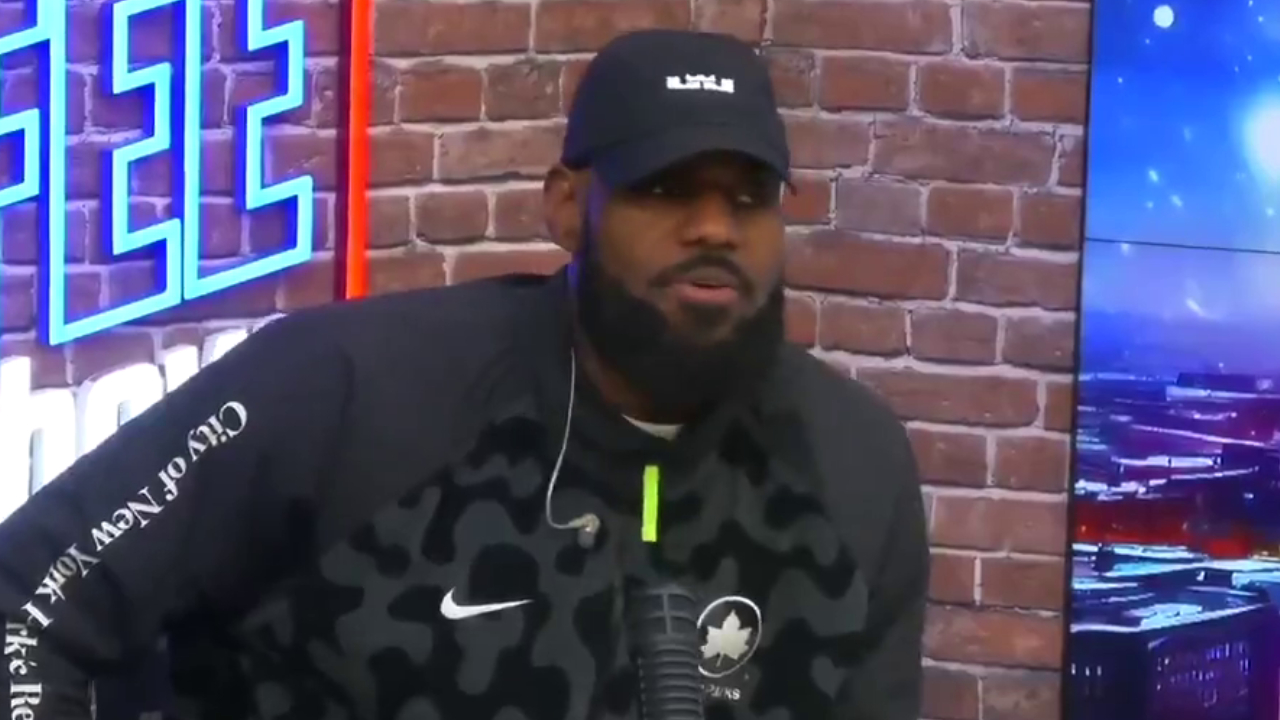 LeBron James wearing a black cap and a Nike jacket is speaking into a microphone in a studio with brick walls and neon lights.