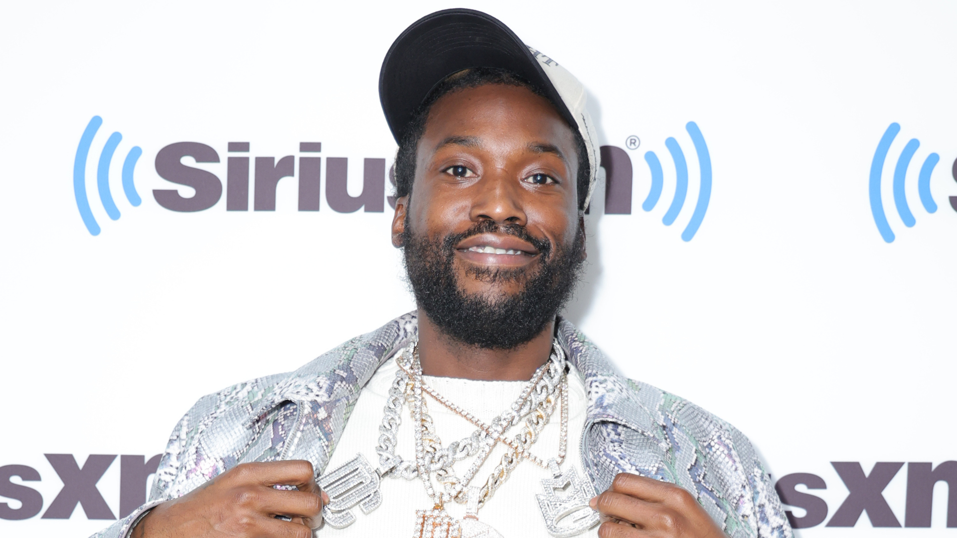 Meek Mill at a SiriusXM event, wearing a patterned jacket, cap, and multiple chains, smiling at the camera.