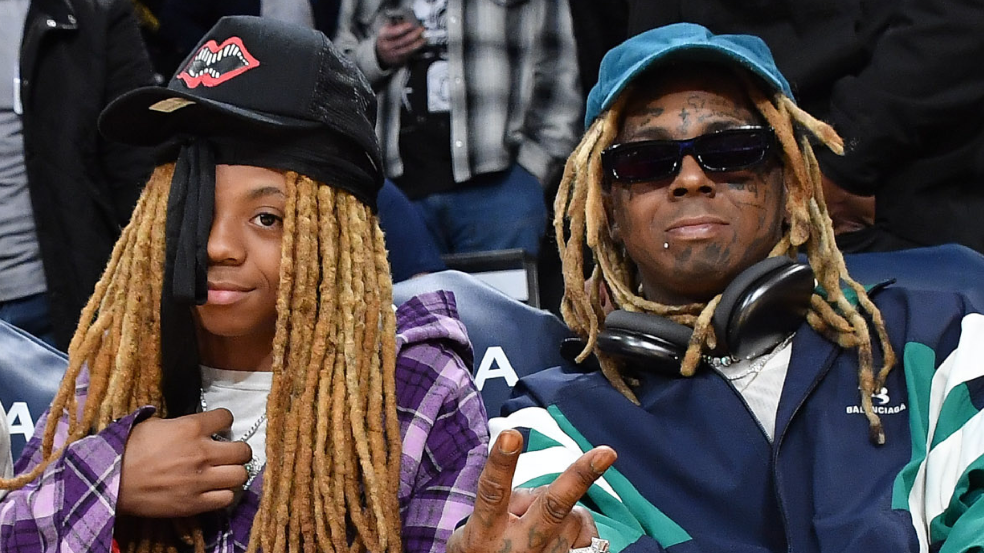 Lil Novi with long hair in a purple plaid shirt and cap sits beside Lil Wayne, who is wearing sunglasses and a blue jacket with headphones.